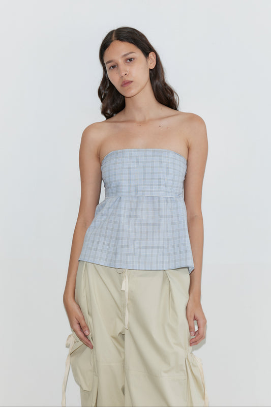 A woman with long dark hair wears the Deiji Studios Flow Top - Powder Check, a strapless cotton top with a removable scarf panel, paired with beige cargo pants. She stands against a plain white background with a neutral expression and relaxed arms.
