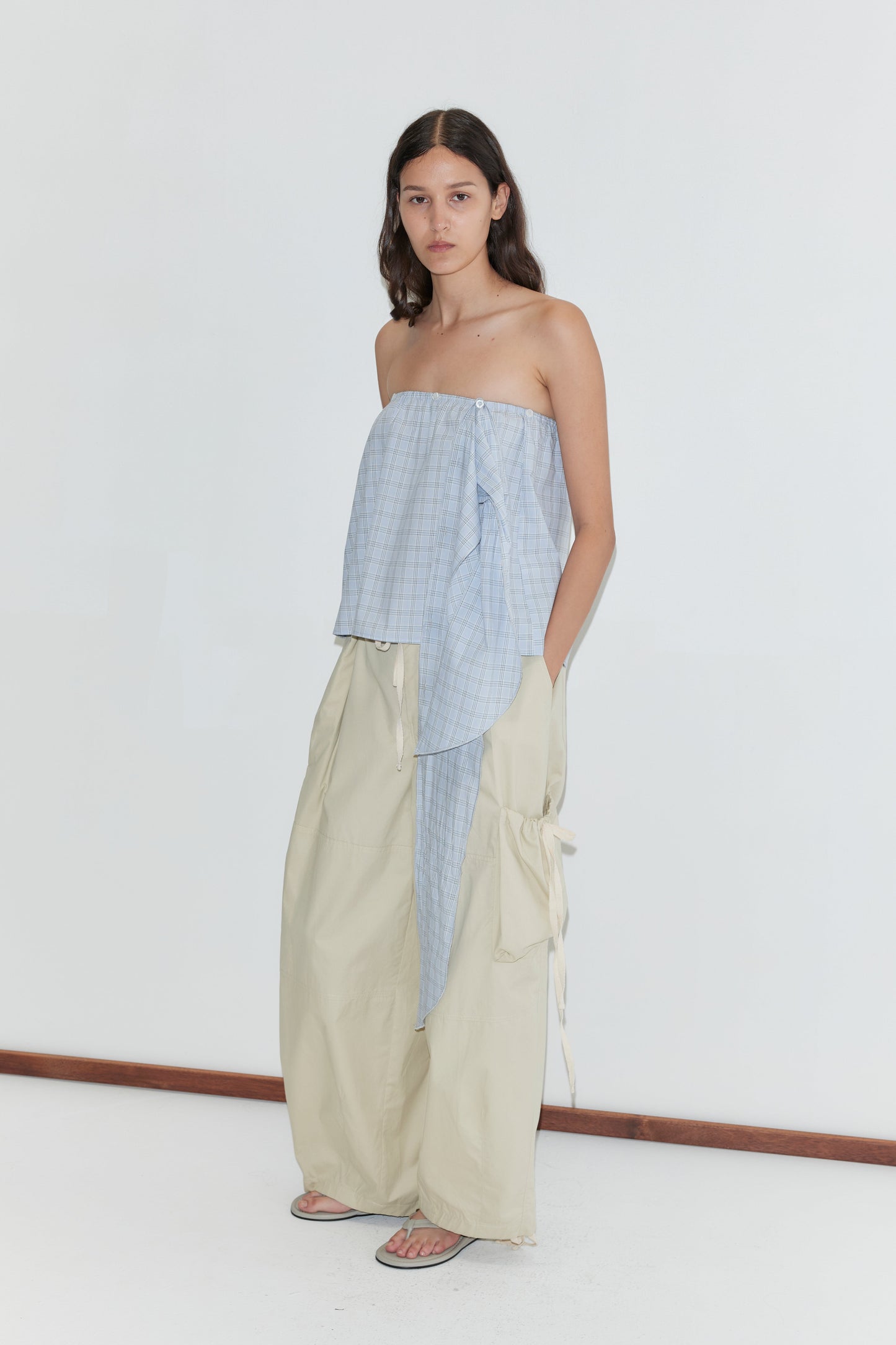 A woman with long dark hair wears a strapless light blue plaid top and Deiji Studios Cargo Pant - Stone, wide-leg beige pants with side ties, standing barefoot against a plain white background.