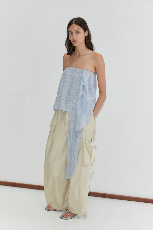 A woman with long dark hair stands against a white background, wearing the Flow Top - Powder Check by Deiji Studios—a strapless cotton top with an asymmetrical blue plaid pattern—paired with loose beige cargo pants and flat sandals.