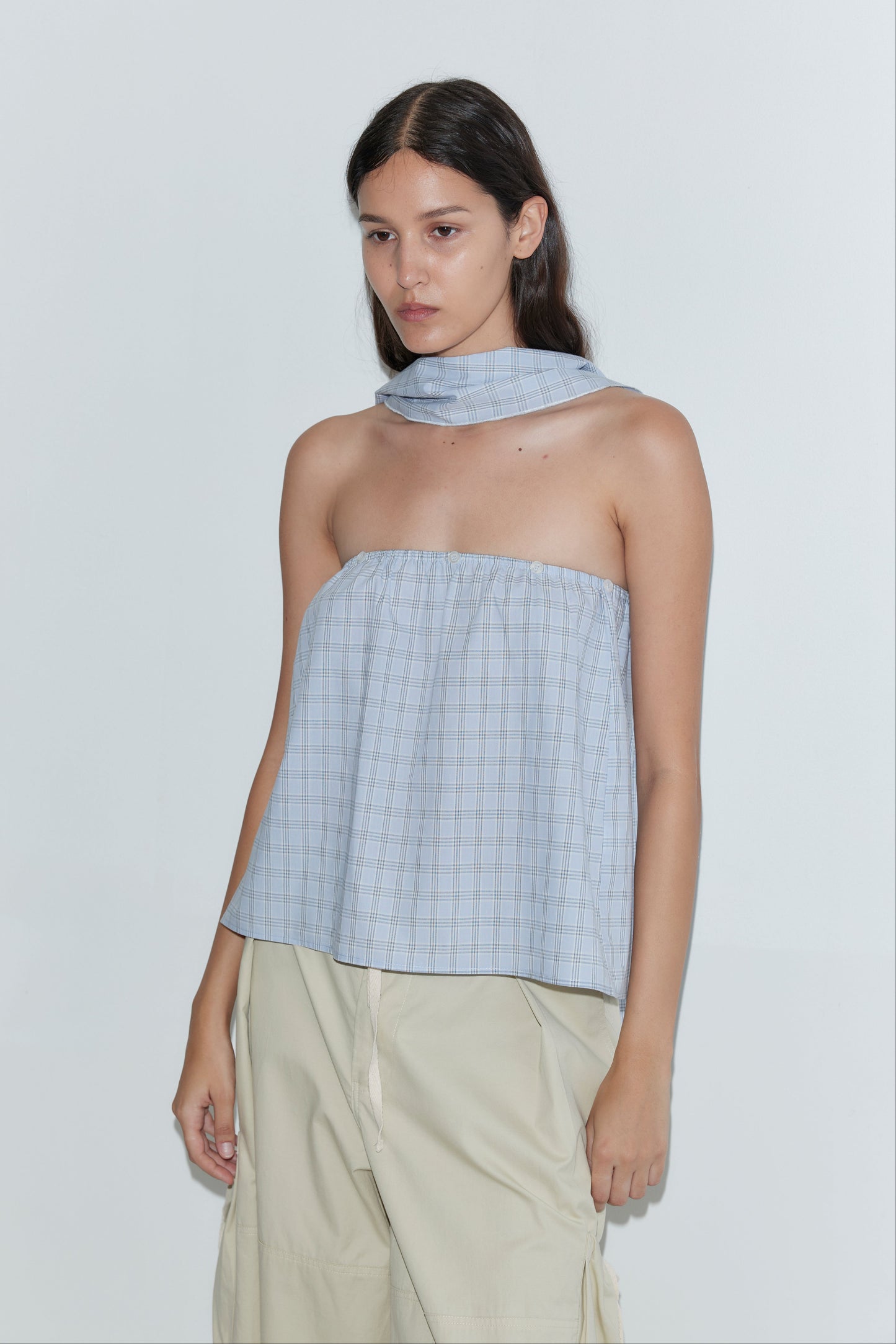 A person with long dark hair wears the Flow Top - Powder Check, a strapless cotton top with a matching removable scarf panel, paired with loose beige pants, standing against a plain white background.