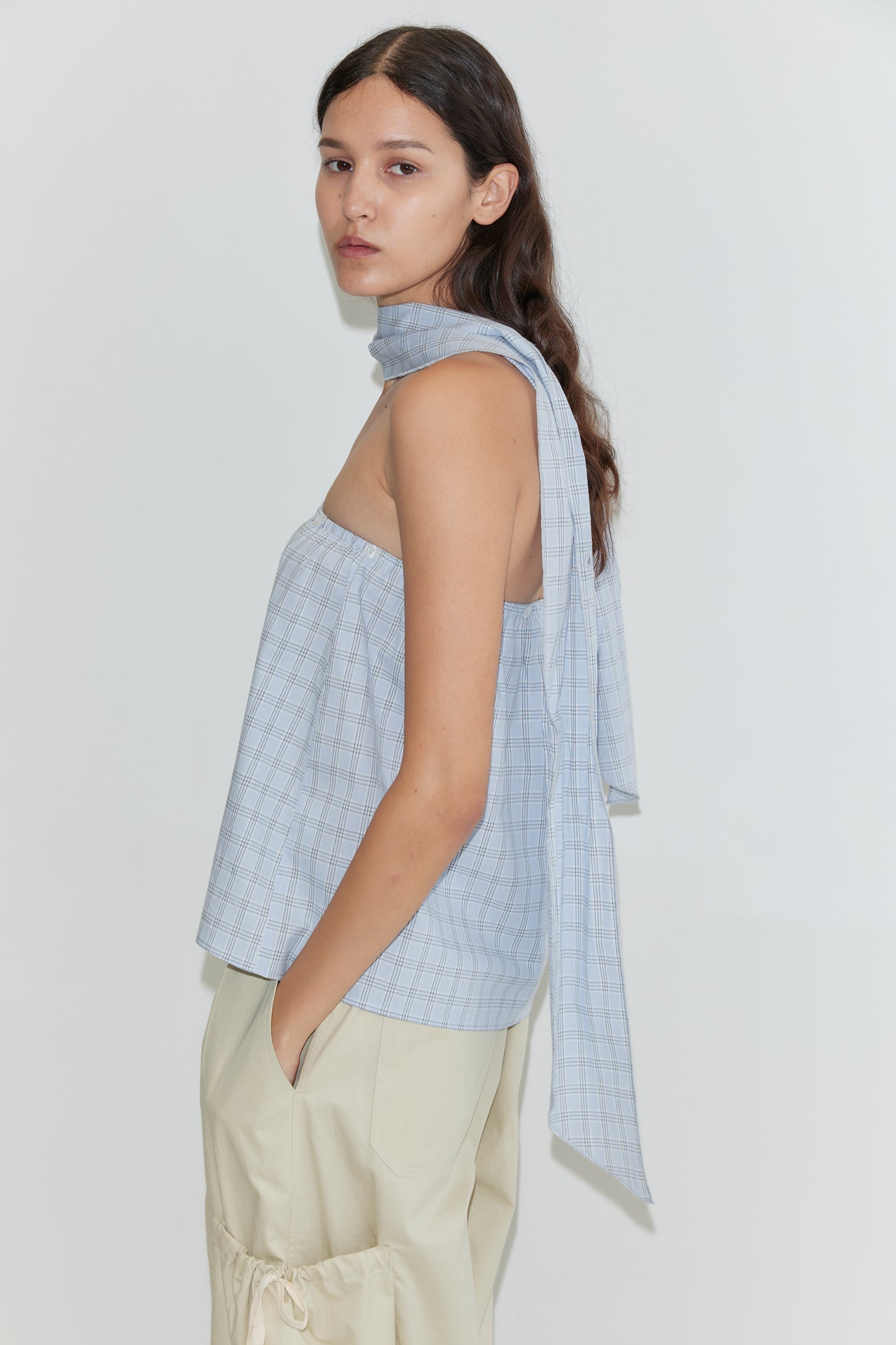 A woman with long brown hair wears the Deiji Studios Flow Top in Powder Check—a light blue checkered, strapless cotton top with a matching removable scarf panel—paired with beige pants. She stands against a plain white background, looking over her shoulder.