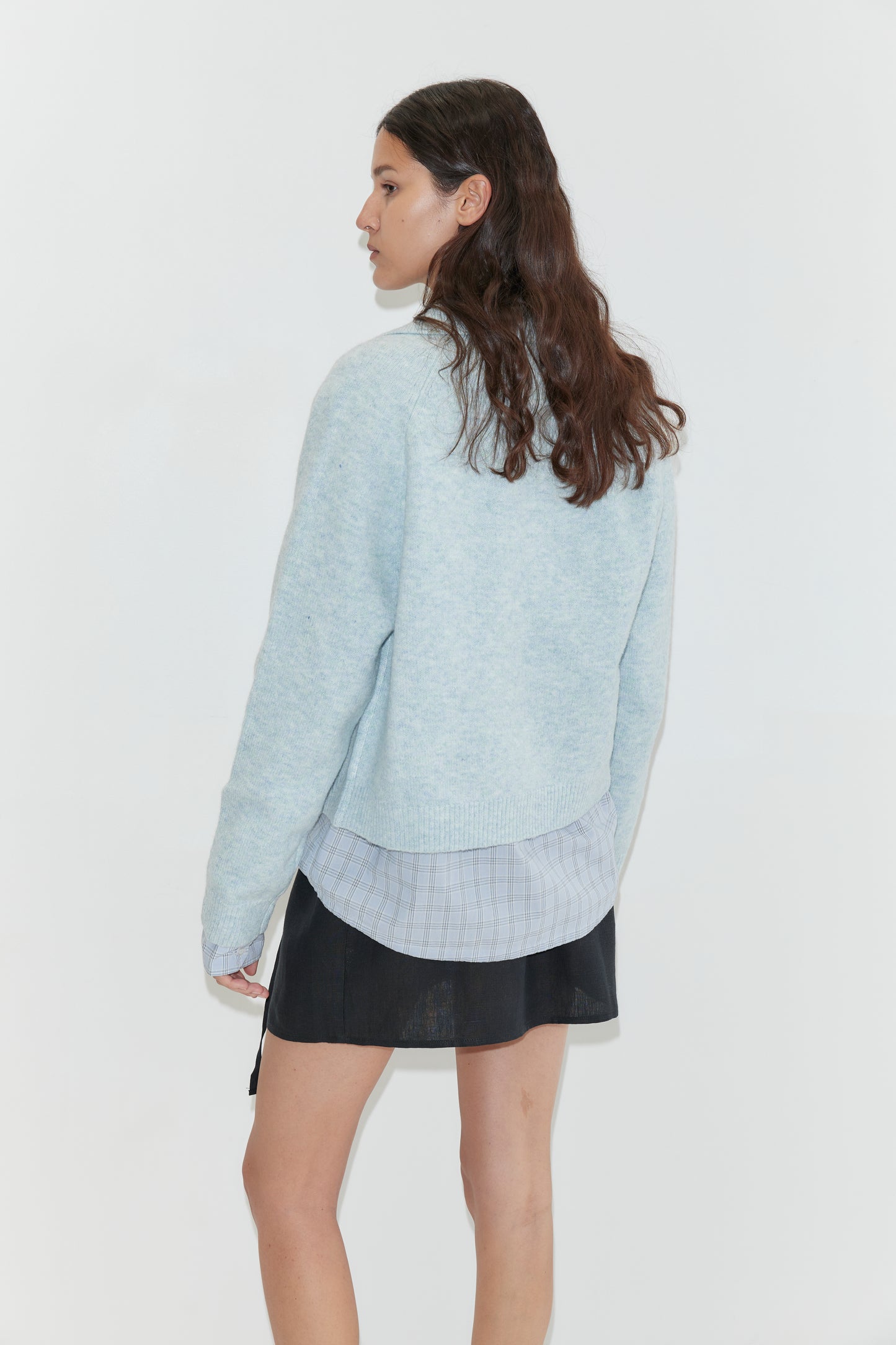 A woman with long brown hair stands facing away, looking to the side against a plain white background. She wears a plaid shirt and black skirt topped with the Deiji Studios Box Cardigan in Young Blue.