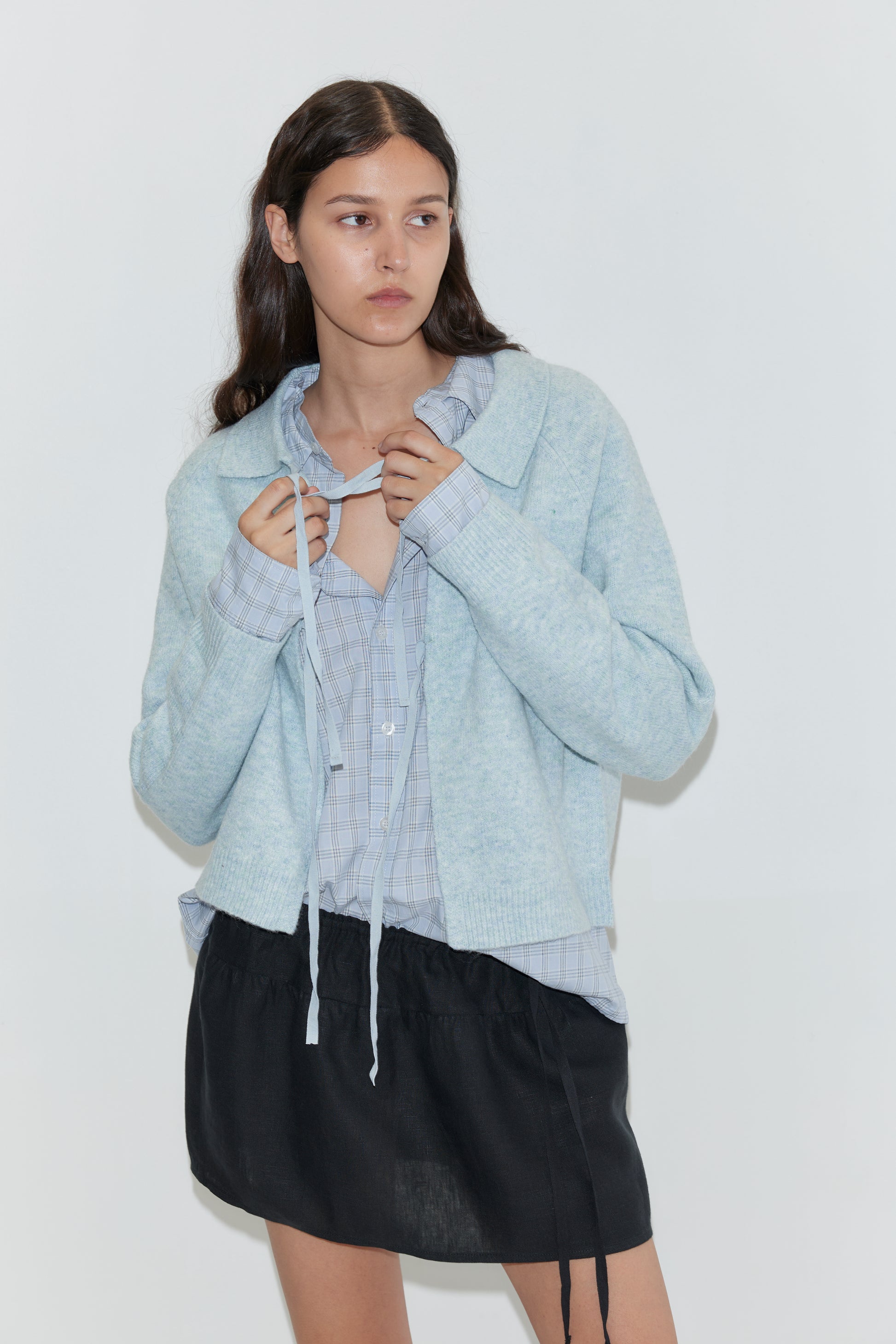 A woman with long dark hair stands against a white background, wearing the Box Cardigan - Young Blue from Deiji Studios over a plaid shirt and black skirt. She holds the cardigan’s drawstrings and gazes slightly to her left.