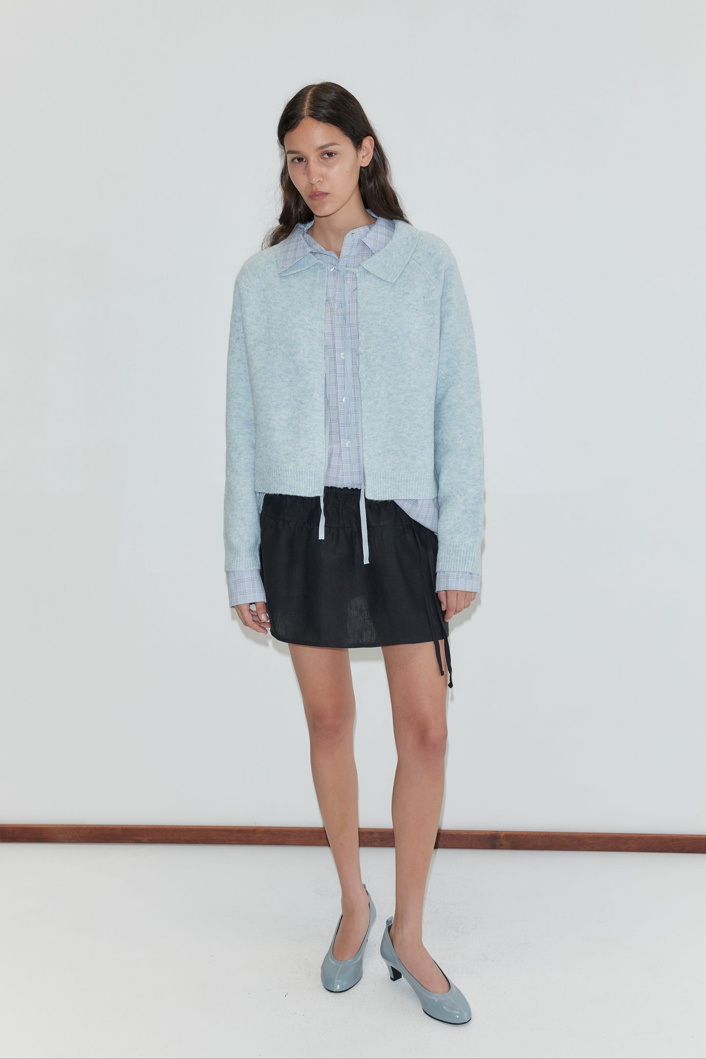 A woman with long, dark hair and a neutral expression stands against a white wall, wearing the Box Cardigan - Young Blue from Deiji Studios over a striped shirt, short black skirt, and matching light blue heels.
