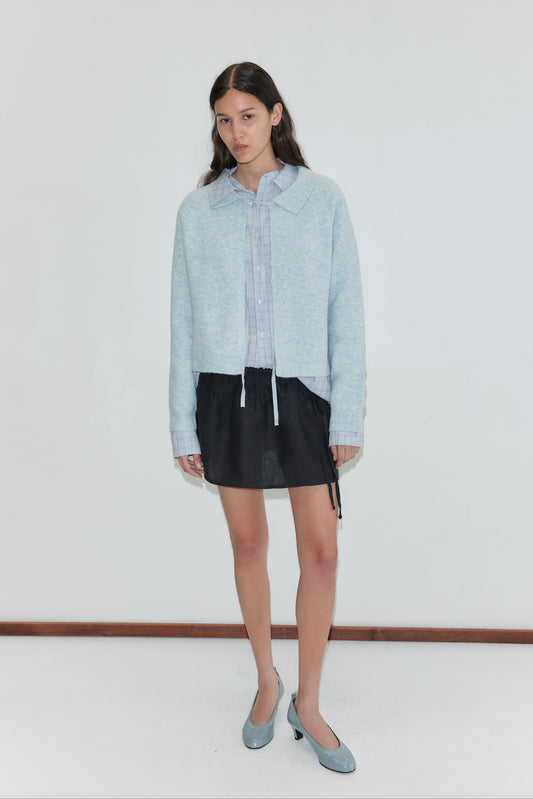 A woman with long, dark hair and a neutral expression stands against a white wall, wearing the Box Cardigan - Young Blue from Deiji Studios over a striped shirt, short black skirt, and matching light blue heels.