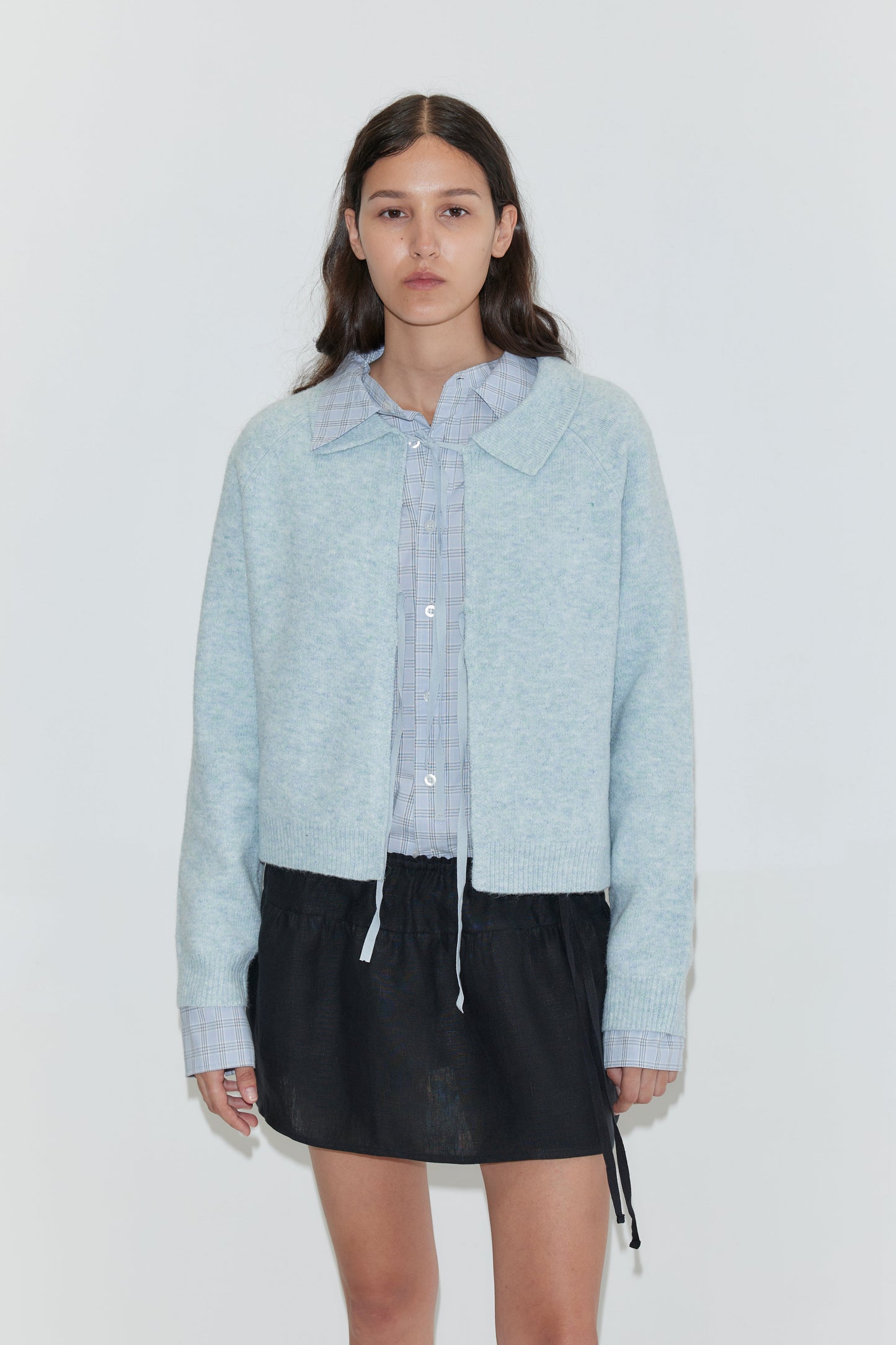 A woman with long brown hair wears the Box Cardigan - Young Blue over a light blue collared shirt and a black mini skirt, standing against a plain white background.