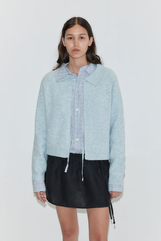 A woman with long brown hair wears the Box Cardigan - Young Blue over a light blue collared shirt and a black mini skirt, standing against a plain white background.
