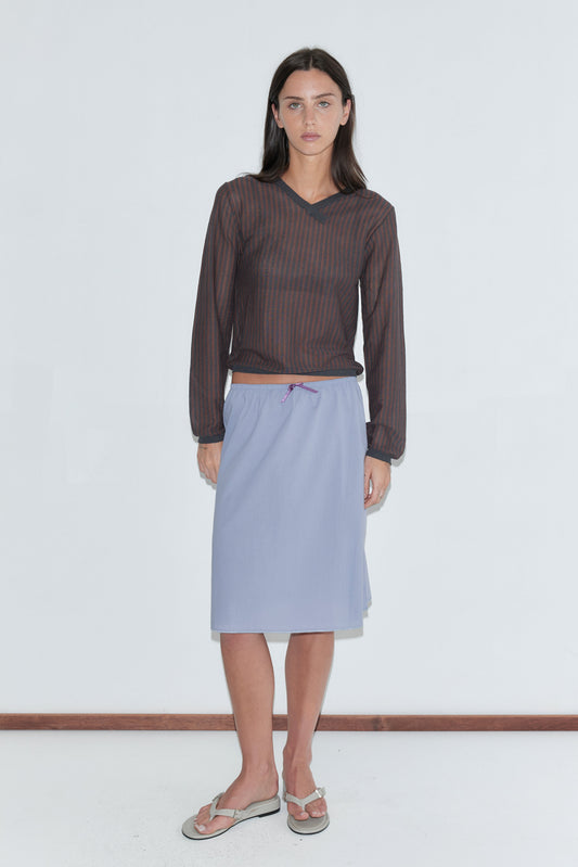 A woman with long dark hair stands against a plain white background wearing the Stripe Longsleeve - Brown Grey Stripe by Deiji Studios, a sheer brown and charcoal striped knit V-neck top, with a periwinkle midi skirt and flip-flops.