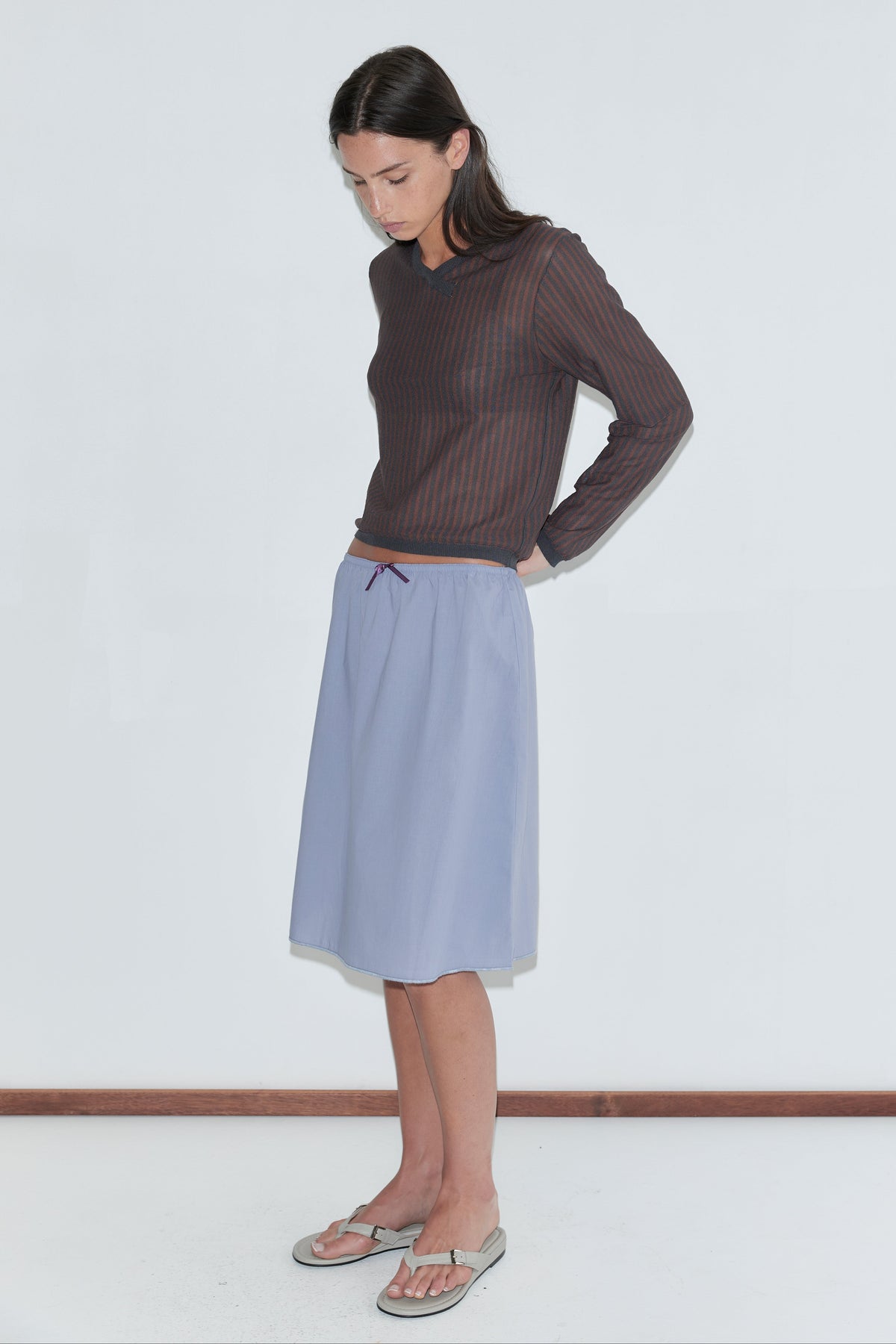 A woman with long dark hair stands in profile against a plain white background wearing the Stripe Longsleeve - Brown Grey Stripe by Deiji Studios, a sheer brown and charcoal striped knit top, with a periwinkle midi skirt and flip-flops.
