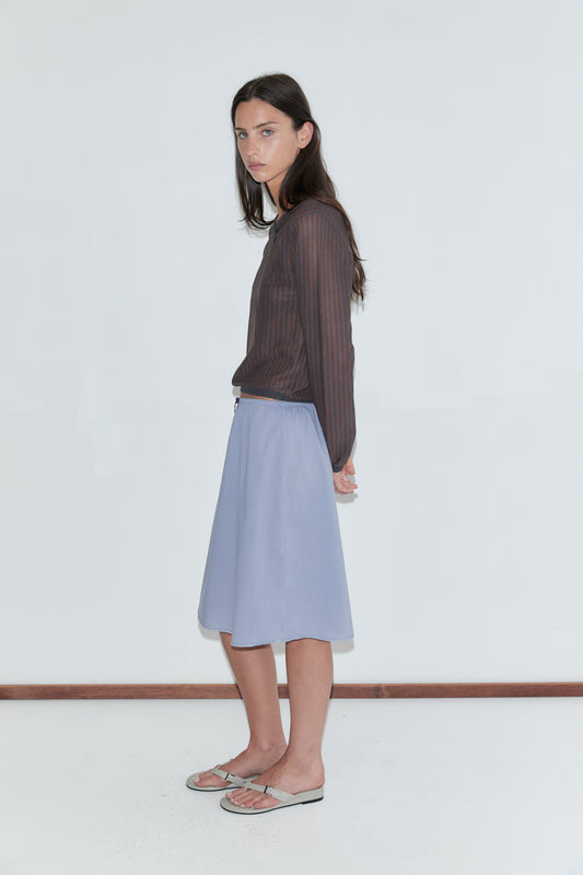 A woman with long dark hair stands in profile against a plain white background wearing a sheer brown striped long-sleeve top and the Basic Skirt - Thistle by Deiji Studios, a periwinkle cotton voile midi skirt with an elasticated waist, and grey flip-flops.