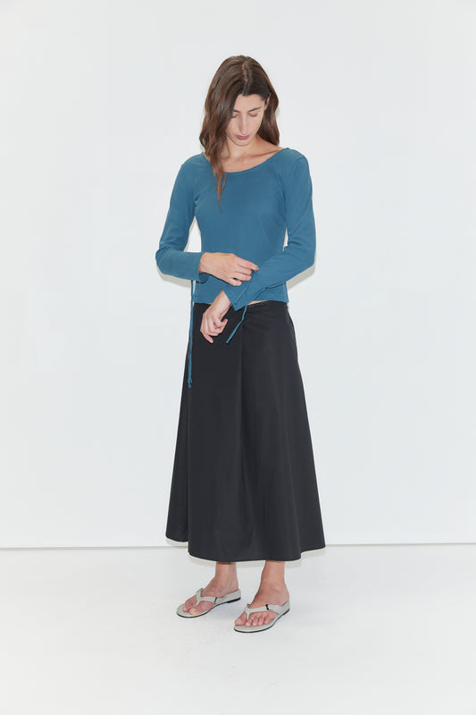 A person stands against a white background, wearing the Cuff Tie Top - Teal from Deiji Studios styled with a black wide-leg skirt and gray flip-flops, looking down with one hand touching their waist.
