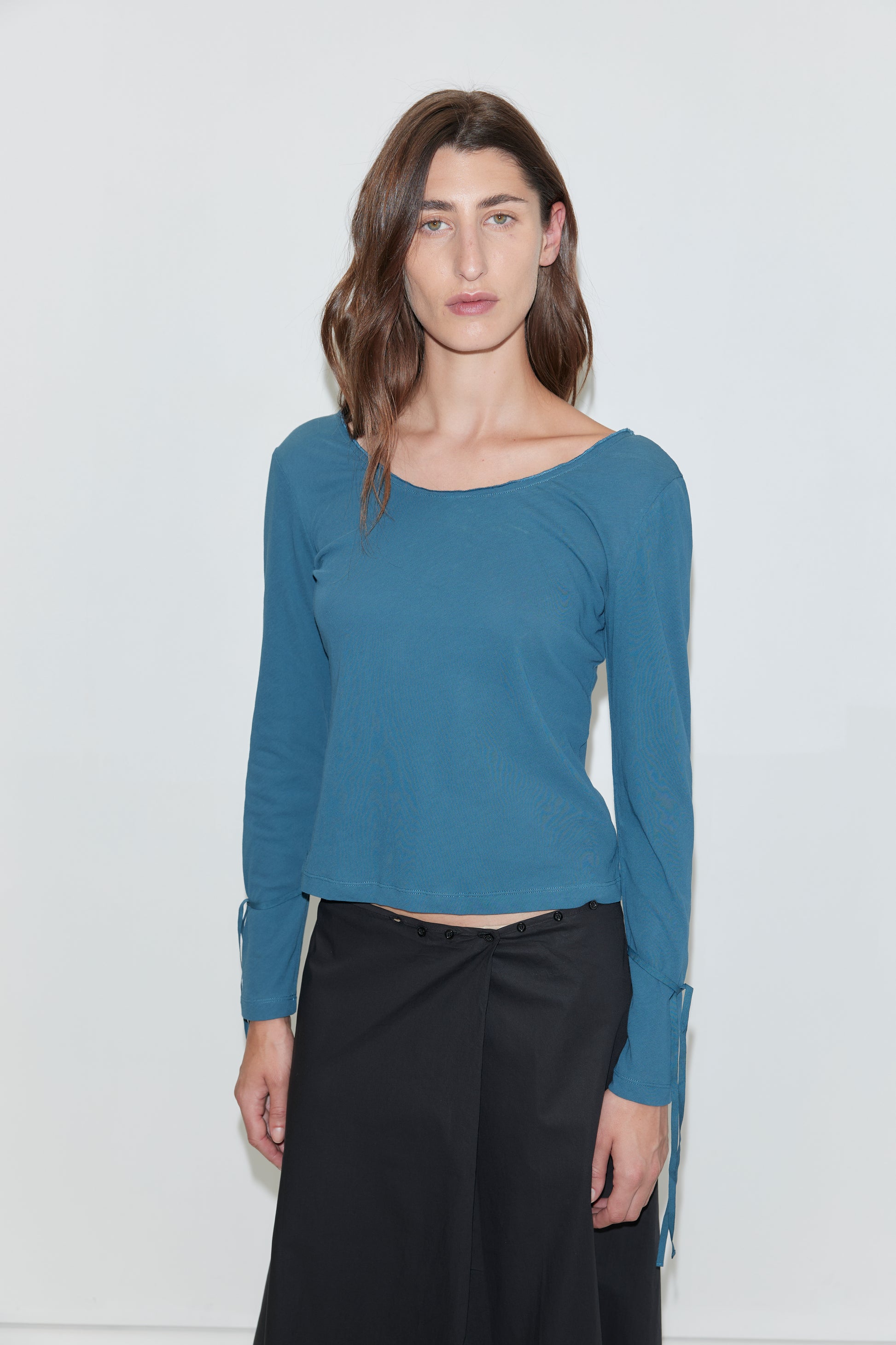 A woman with wavy brown hair stands against a white background, wearing the Cuff Tie Top - Teal from Deiji Studios, featuring long sleeves with small side ties at the cuffs, paired with a black button-front skirt. She faces forward with a neutral expression.