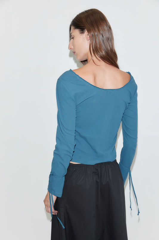 A person with long brown hair, wearing a teal cuff tie top from Deiji Studios and a black skirt, is standing against a plain white background, facing away and slightly to the side.