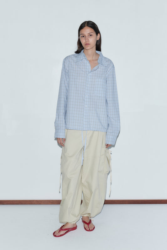 A person with long dark hair wears a Stand Shirt - Powder Check and wide beige cargo pants, paired with red flip-flops, standing against a plain white wall.