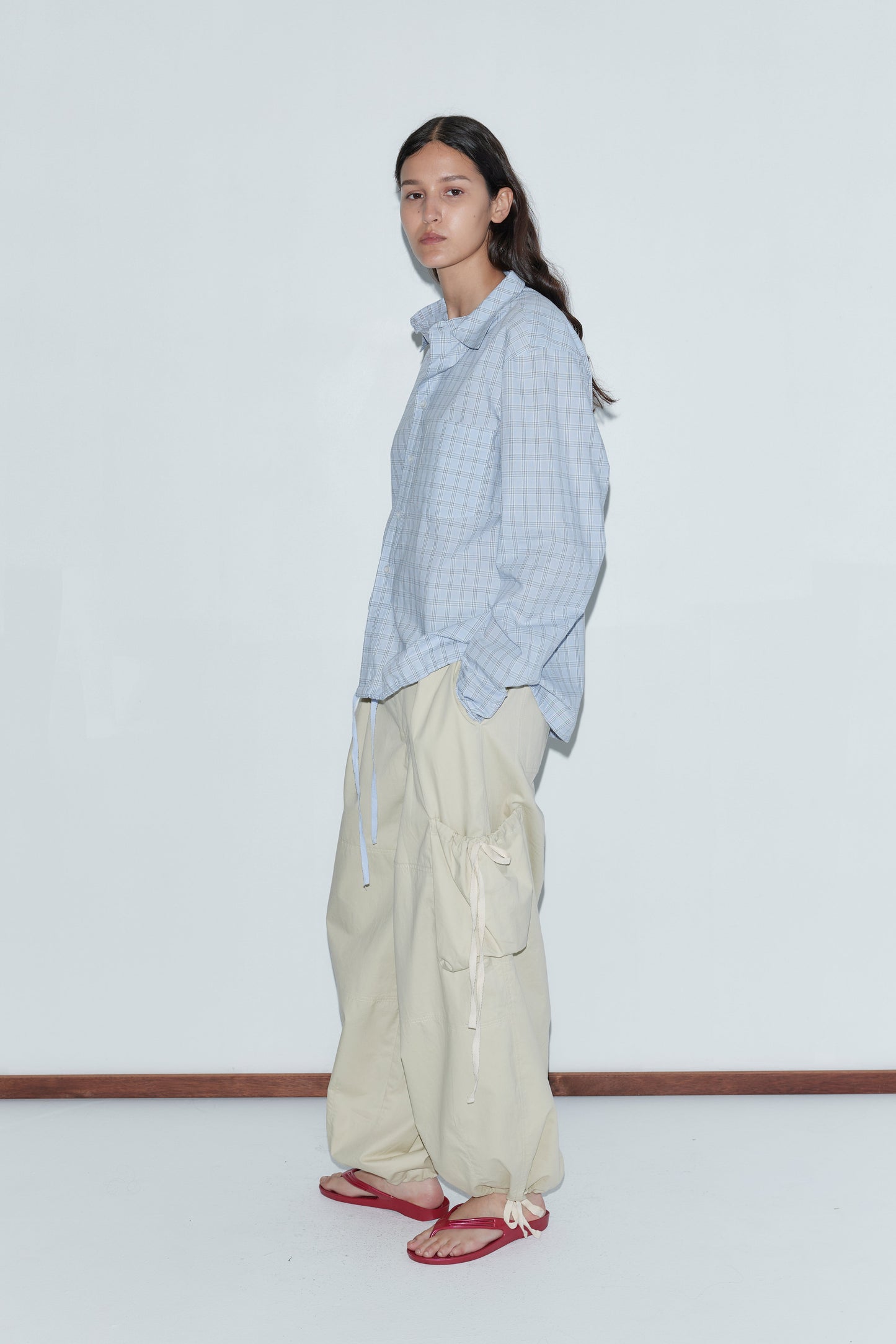 A person stands against a plain white wall wearing the Stand Shirt - Powder Check, an oversized cotton shirt with a light blue check pattern, paired with beige cargo pants and red flip-flops, embodying classic Deiji Studios style.