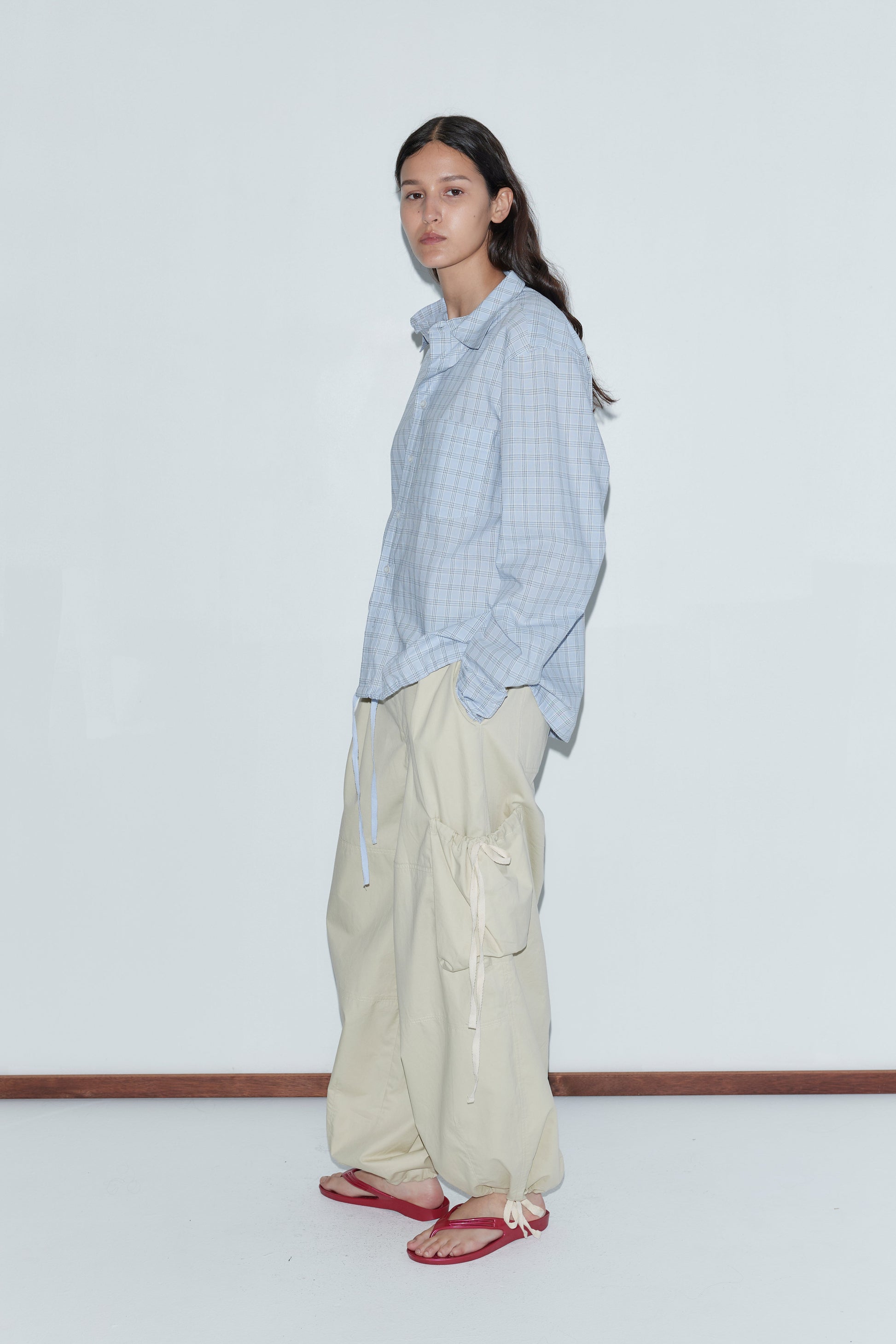 A person stands against a plain white wall wearing the Stand Shirt - Powder Check, an oversized cotton shirt with a light blue check pattern, paired with beige cargo pants and red flip-flops, embodying classic Deiji Studios style.