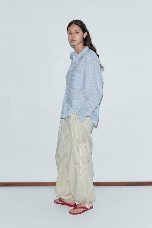 A person stands against a plain white wall wearing the Stand Shirt - Powder Check, an oversized cotton shirt with a light blue check pattern, paired with beige cargo pants and red flip-flops, embodying classic Deiji Studios style.