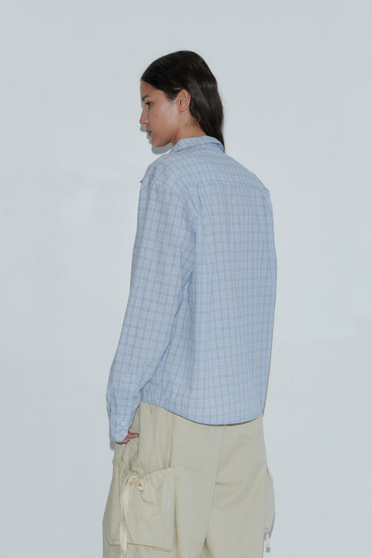 A person with long dark hair, wearing the Stand Shirt - Powder Check and beige cargo pants, stands facing away and slightly to the side against a plain light background.