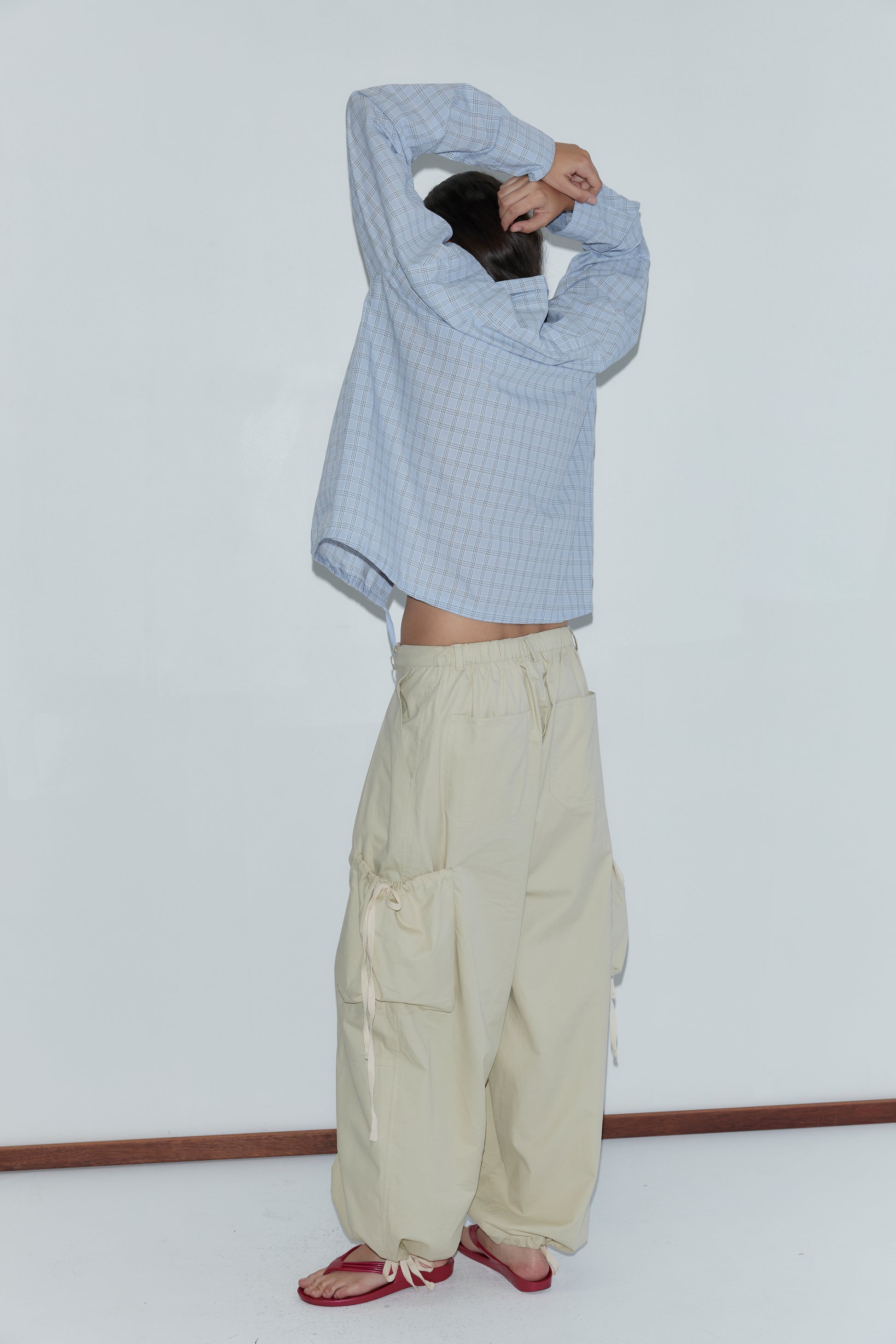 A person faces away, lifting a light blue plaid shirt over their head. They wear Deiji Studios Cargo Pant in Stone with side ties and red sandals, standing against a plain white wall and wooden floor.