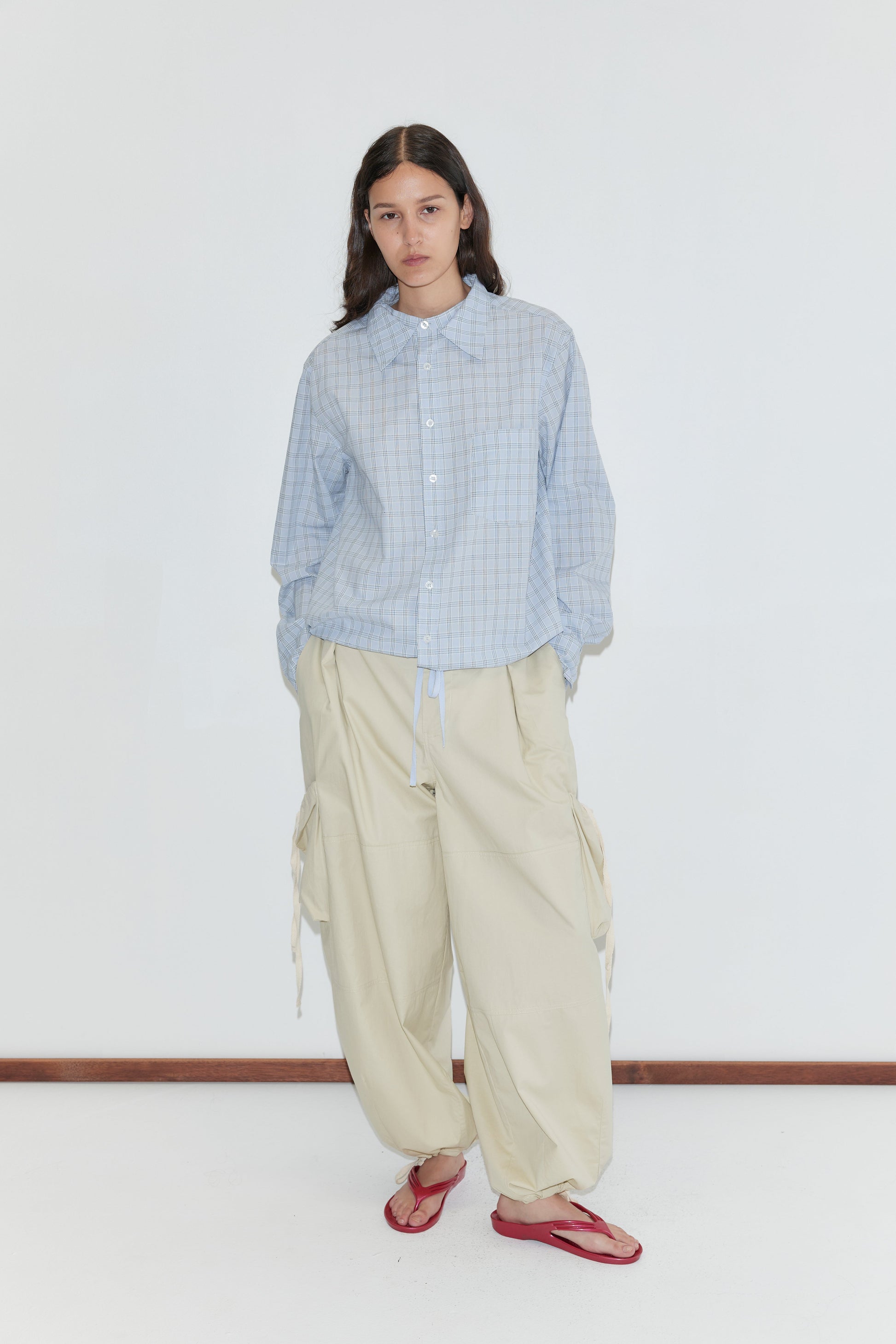 A person with long dark hair and a neutral expression stands against a plain white background, wearing the Stand Shirt - Powder Check by Deiji Studios, loose beige cargo pants with drawstrings, and red flip-flops.
