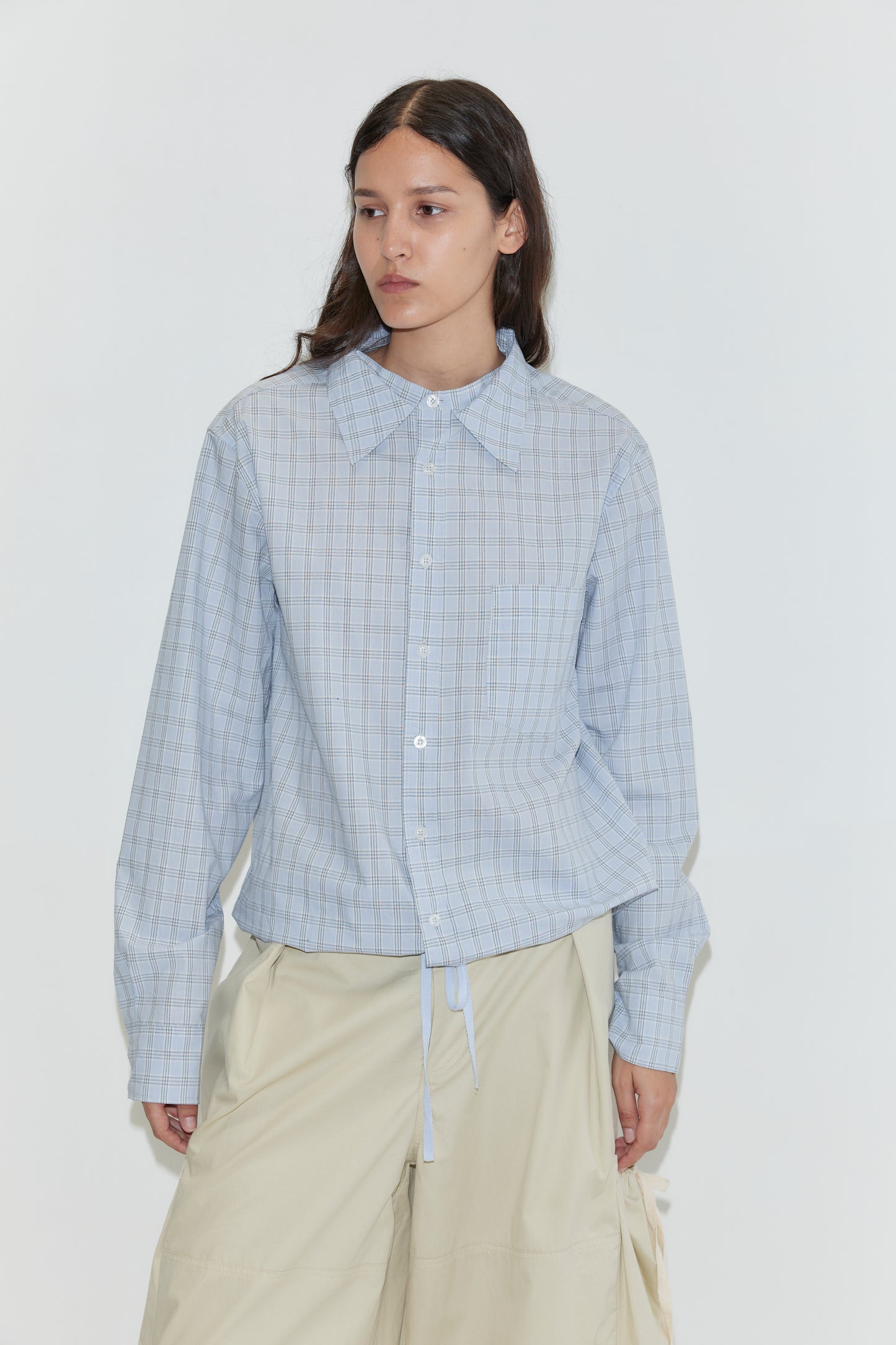 A person with long dark hair wears the Stand Shirt - Powder Check, an oversized cotton shirt with a drawstring hem, inspired by Deiji Studios, paired with cream wide-leg pants against a plain white background.