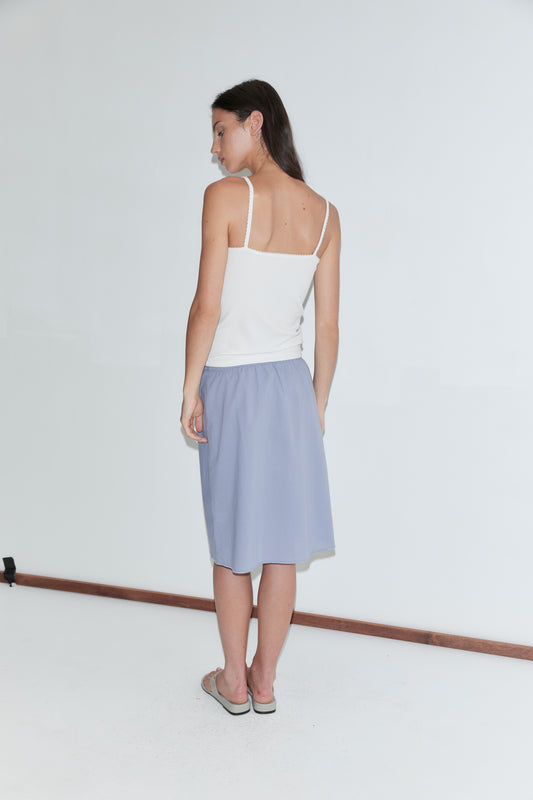 A woman with long dark hair stands with her back to the camera against a plain white background, wearing a white scallop-strap tank and the Basic Skirt - Thistle by Deiji Studios, a periwinkle cotton voile midi skirt, with grey flip-flops.