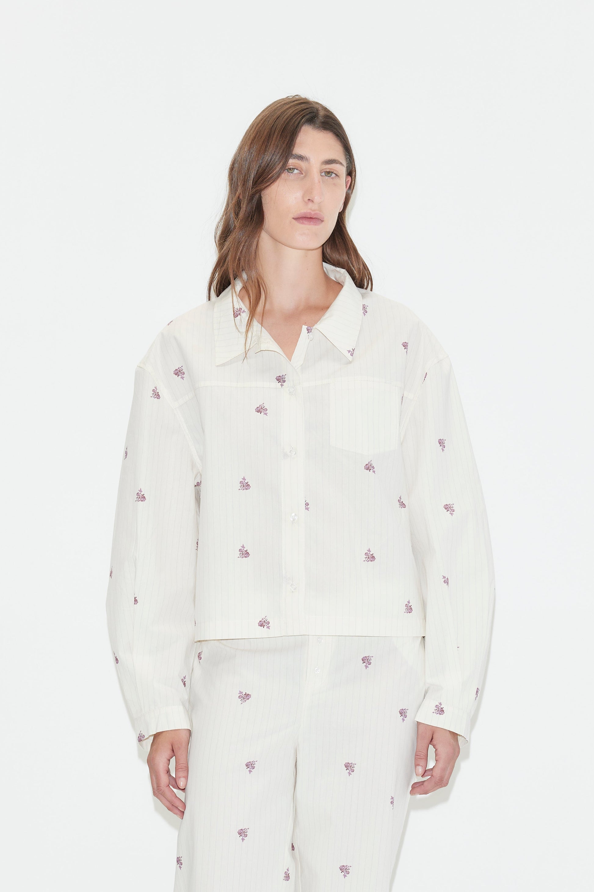 A person with long brown hair wears a loose-fitting white Boxy Shirt - Floral Stripe pajama set, featuring an oversized shirt adorned with small purple floral stripes, standing against a plain white background.