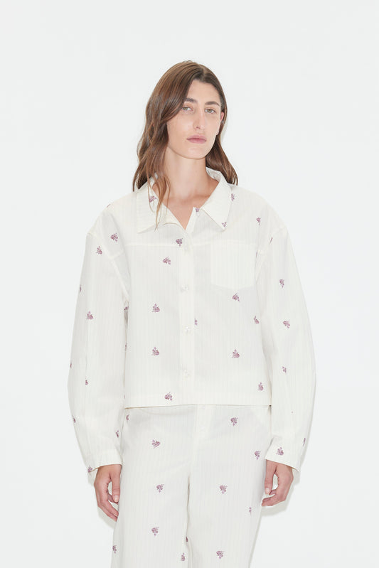 A person with long brown hair wears a loose-fitting white Boxy Shirt - Floral Stripe pajama set, featuring an oversized shirt adorned with small purple floral stripes, standing against a plain white background.