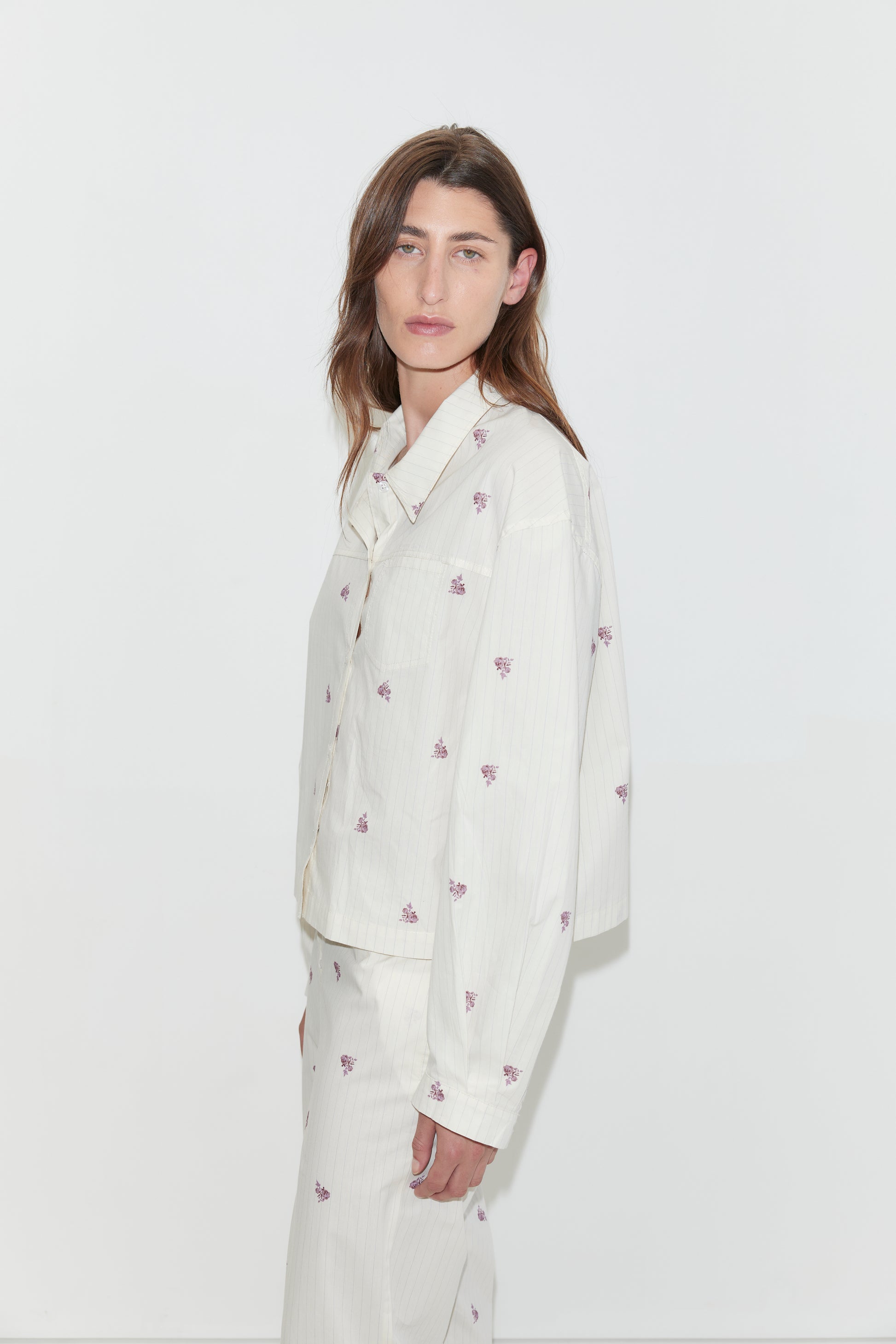 A person with long brown hair, wearing an oversized light-colored organic cotton Boxy Shirt - Floral Stripe and matching pants with small purple floral prints, stands against a plain white background, looking slightly toward the camera.
