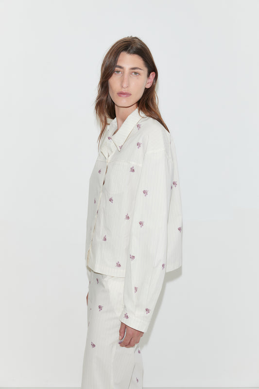 A person with long brown hair, wearing an oversized light-colored organic cotton Boxy Shirt - Floral Stripe and matching pants with small purple floral prints, stands against a plain white background, looking slightly toward the camera.