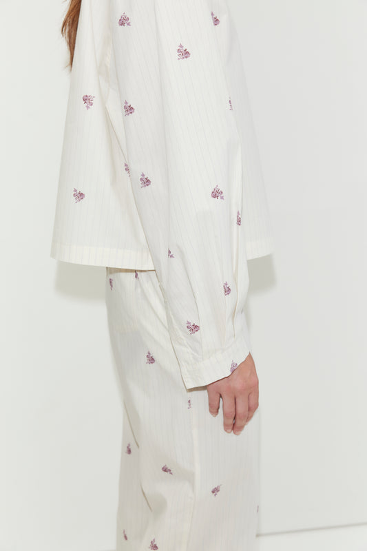 A person stands sideways against a plain white background, wearing the Boxy Long Set - Floral Stripe. This cream-colored pajama set features thin stripes and small purple floral prints, and is crafted from cotton poplin for a relaxed, oversized fit with a matching shirt and bottoms.
