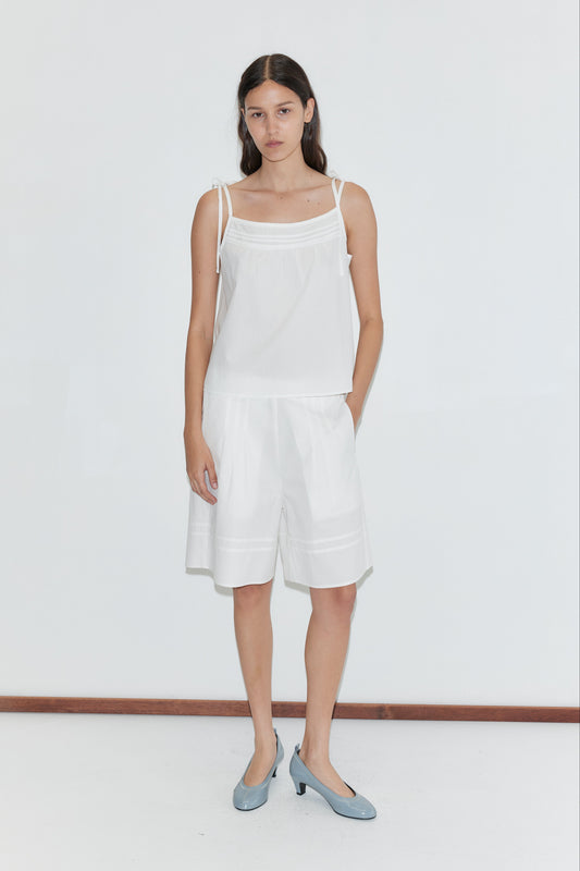 A woman with long dark hair and a neutral expression stands against a white wall, wearing the Tied Cami - White by Deiji Studios, paired with loose white knee-length shorts and light blue low-heeled shoes.