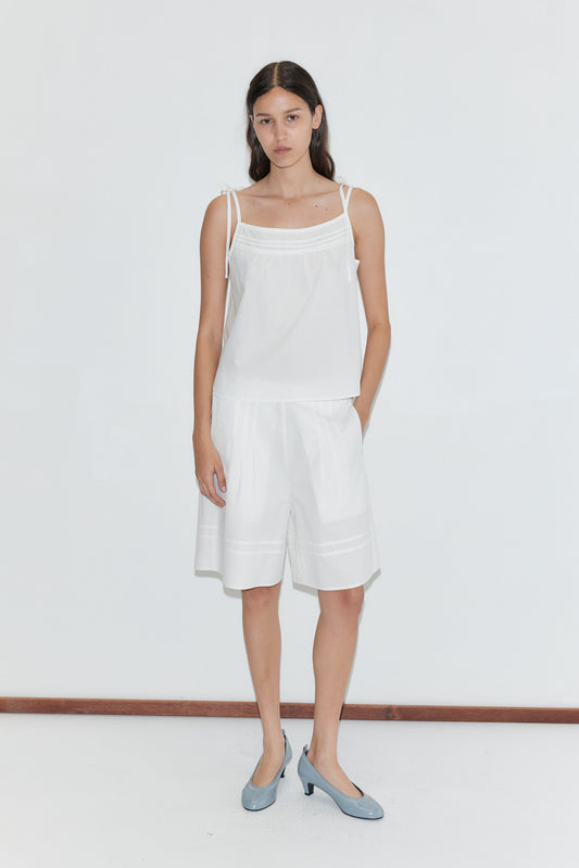 A woman with long dark hair stands against a plain white wall wearing a sleeveless white top and the Pintuck Short - White by Deiji Studios, featuring pintuck detailing. She pairs them with light blue low-heeled shoes and keeps one hand in her pocket, neutral-faced.