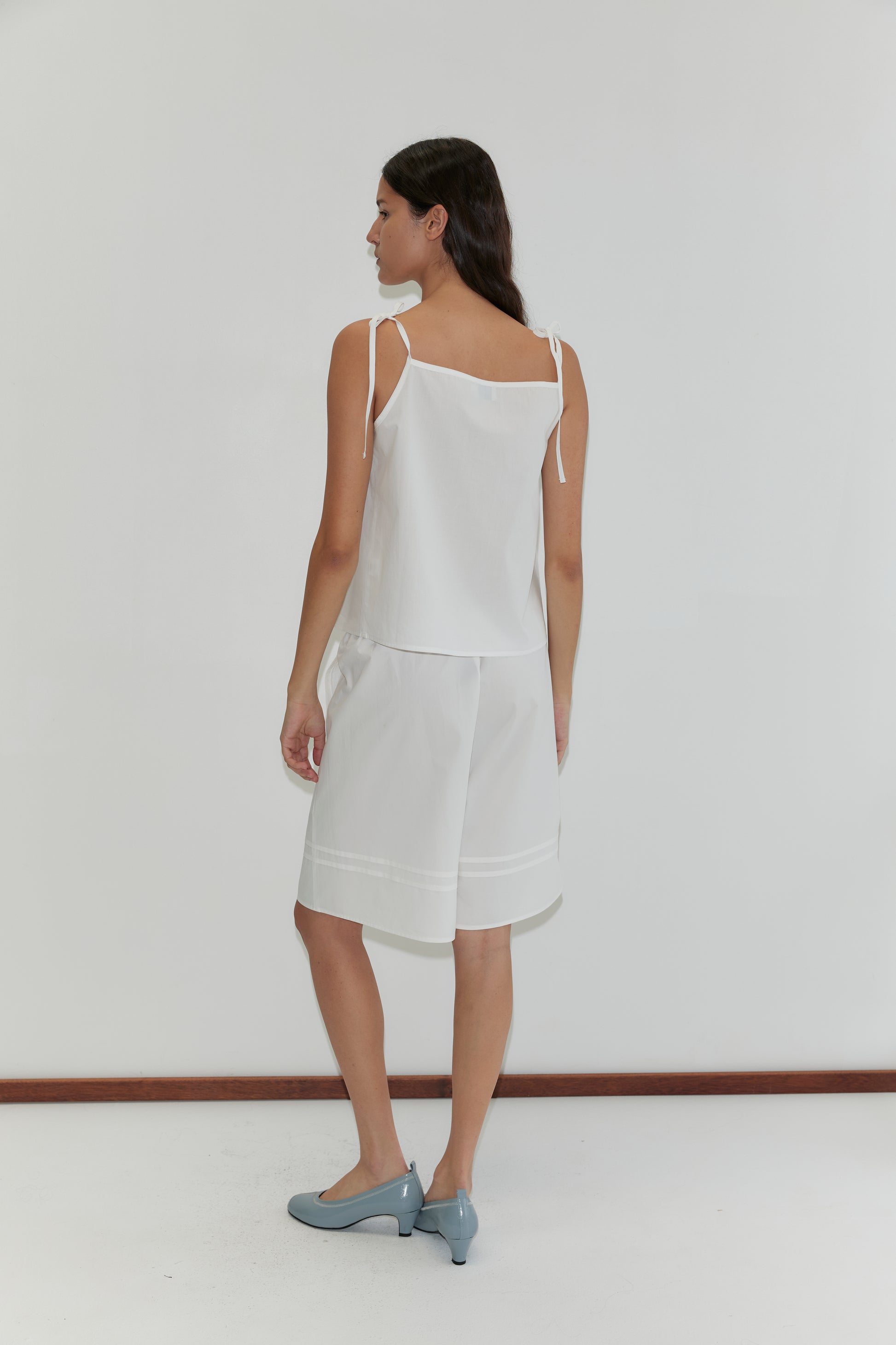 A woman with long brown hair wears the Deiji Studios Tied Cami - White and loose white knee-length shorts, paired with light blue low-heeled shoes. She stands facing away against a plain white background.