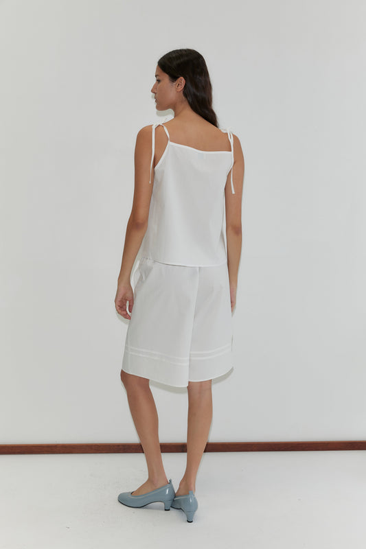 A woman with long dark hair, seen from behind, wears the Pintuck Short - White by Deiji Studios and a sleeveless top with tied shoulder straps, paired with light blue low-heeled shoes against a plain background.