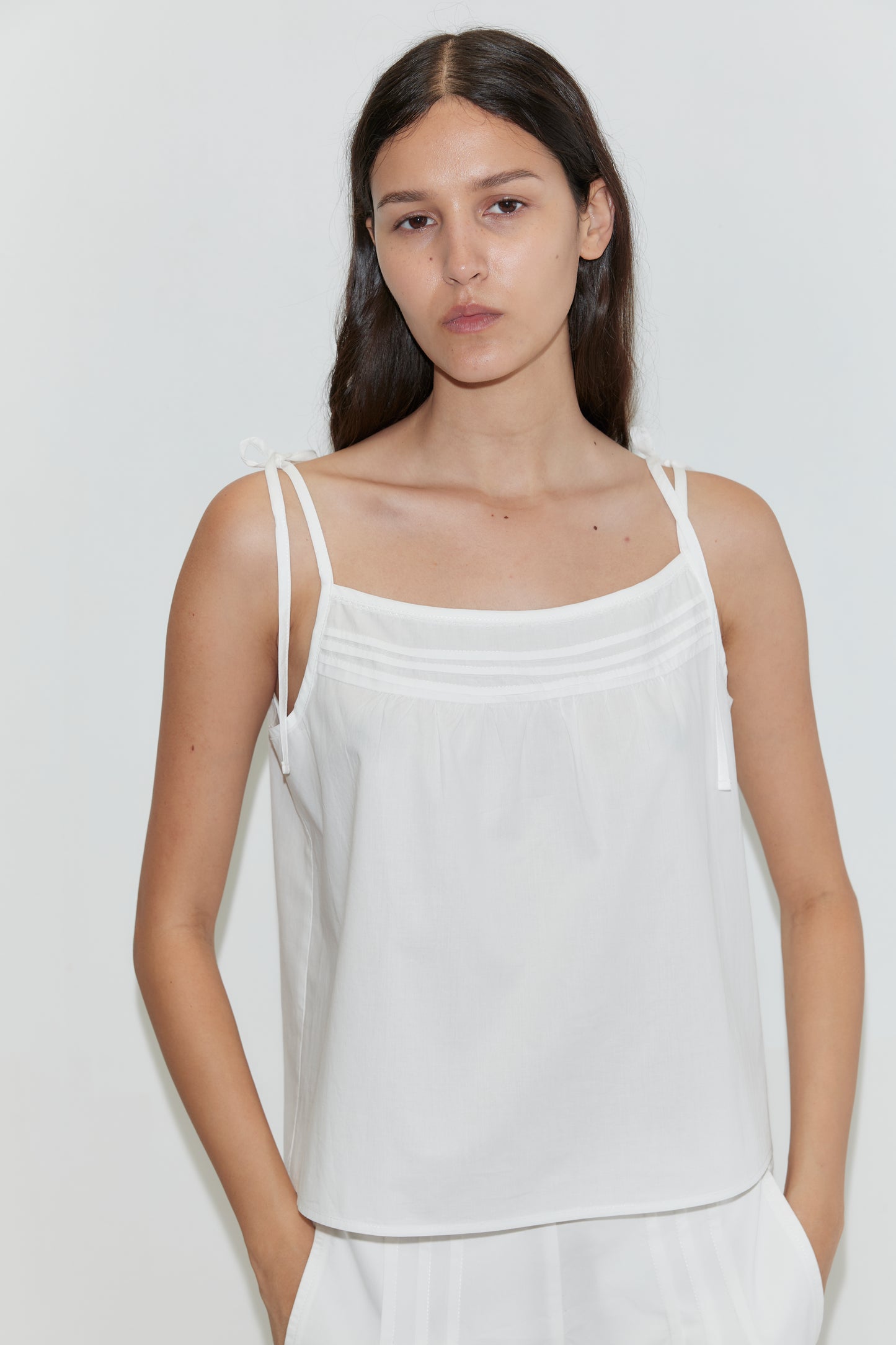 A woman with long, dark hair wears the Tied Cami - White with thin straps, standing before a plain white background and gazing neutrally at the camera.