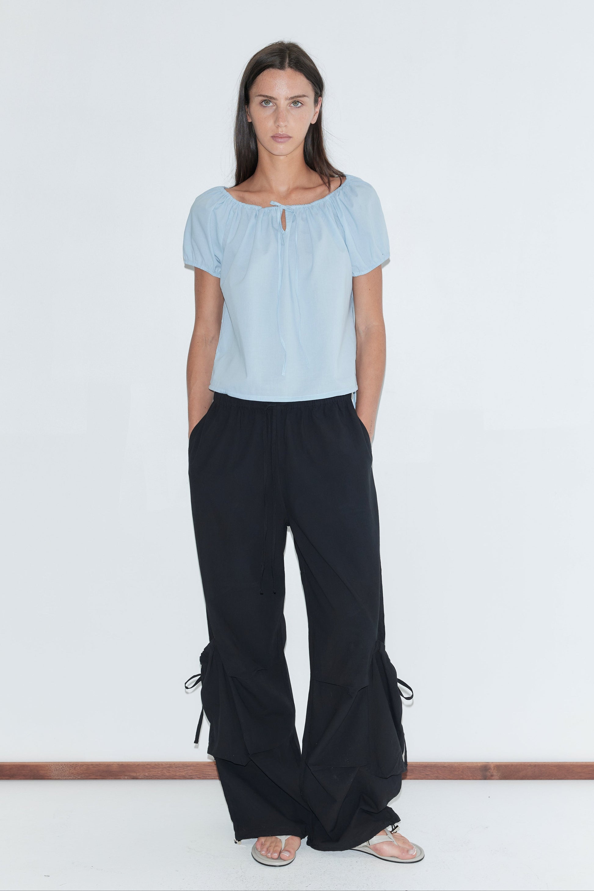 A woman with long dark hair stands against a plain white background wearing the Tie Top - Blue by Deiji Studios, a pale blue top with a gathered neckline and puff sleeves, with black wide-leg trousers and flip-flops.