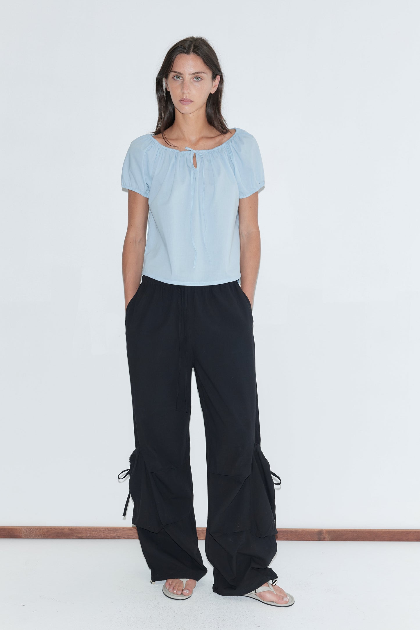A woman with long dark hair stands against a plain white background wearing the Drawcord Pocket Pant - Black by Deiji Studios, black wide-leg trousers with drawcord-gathered lower legs, with a pale blue tie top and flip-flops.