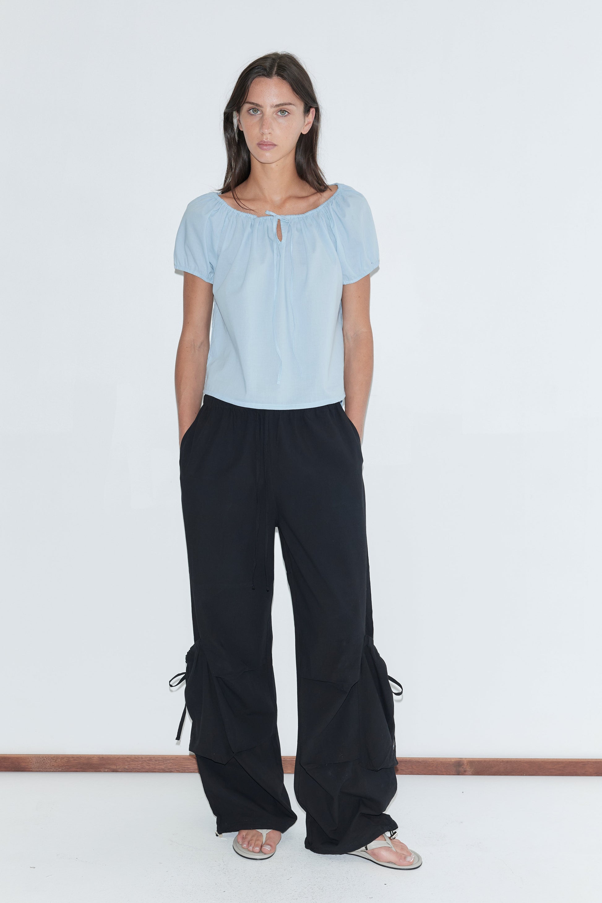 A woman with long dark hair stands against a plain white background wearing the Drawcord Pocket Pant - Black by Deiji Studios, black wide-leg trousers with drawcord-gathered lower legs, with a pale blue tie top and flip-flops.