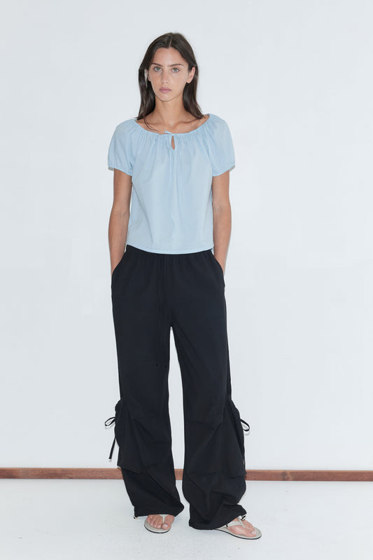 A woman with long dark hair stands against a plain white background wearing the Drawcord Pocket Pant - Black by Deiji Studios, black wide-leg trousers with drawcord-gathered lower legs, with a pale blue tie top and flip-flops.