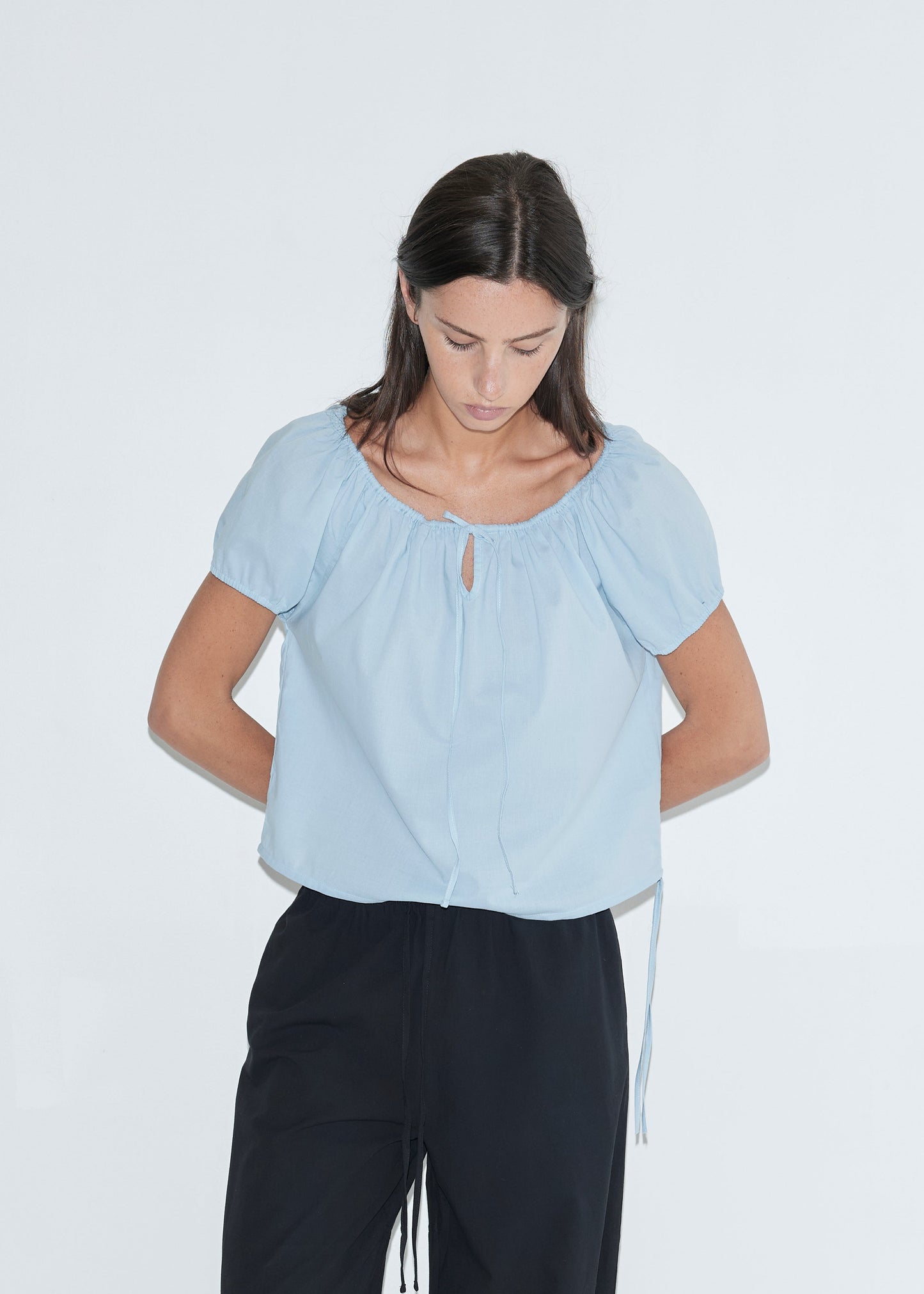 A woman with long dark hair stands against a plain white background looking down, wearing the Tie Top - Blue by Deiji Studios, a pale blue top with a gathered neckline, keyhole drawstring tie and puff sleeves, with black trousers.
