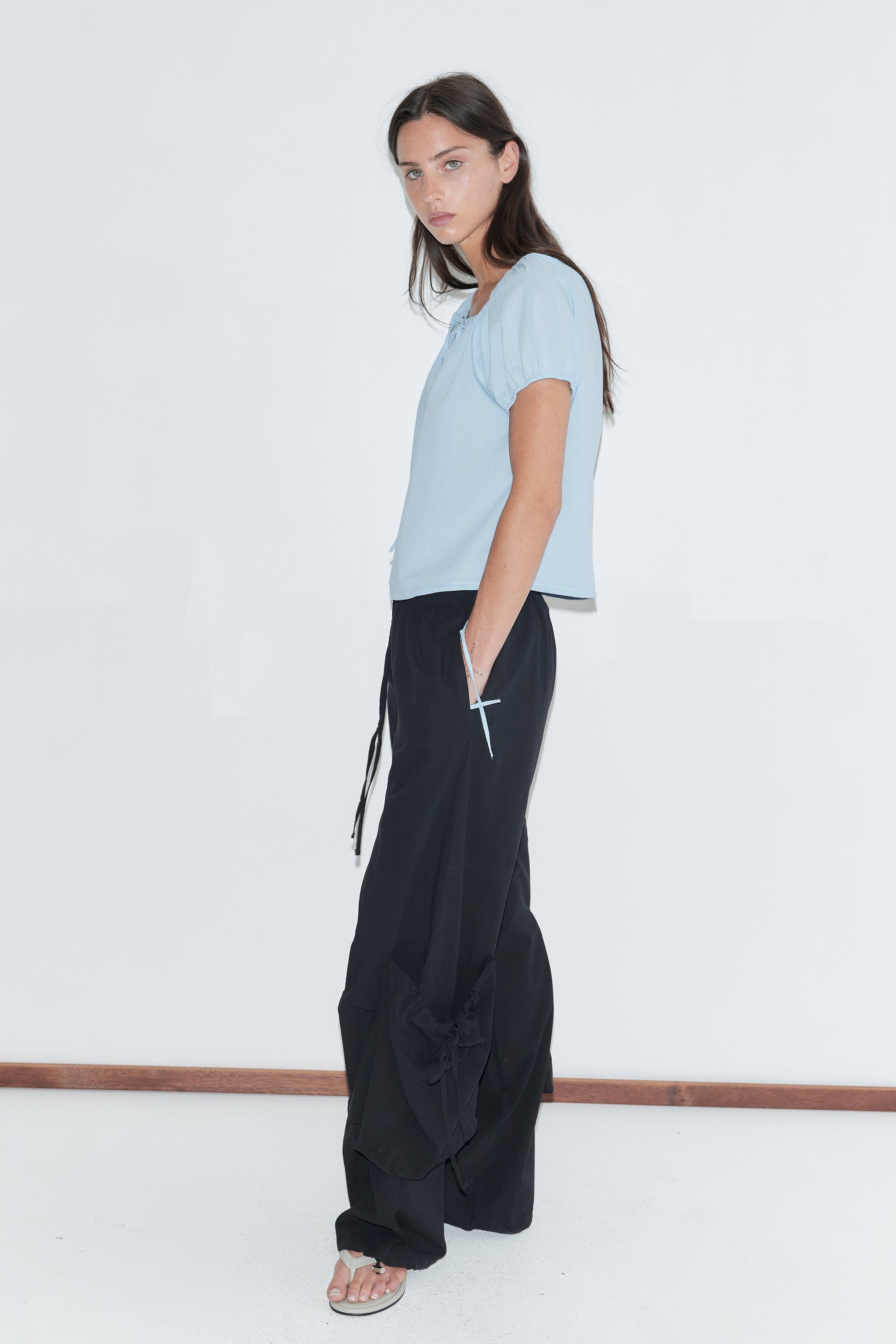 A woman with long dark hair stands in profile against a plain white background wearing the Tie Top - Blue by Deiji Studios, a pale blue top with a gathered neckline, with black wide-leg trousers and flip-flops.