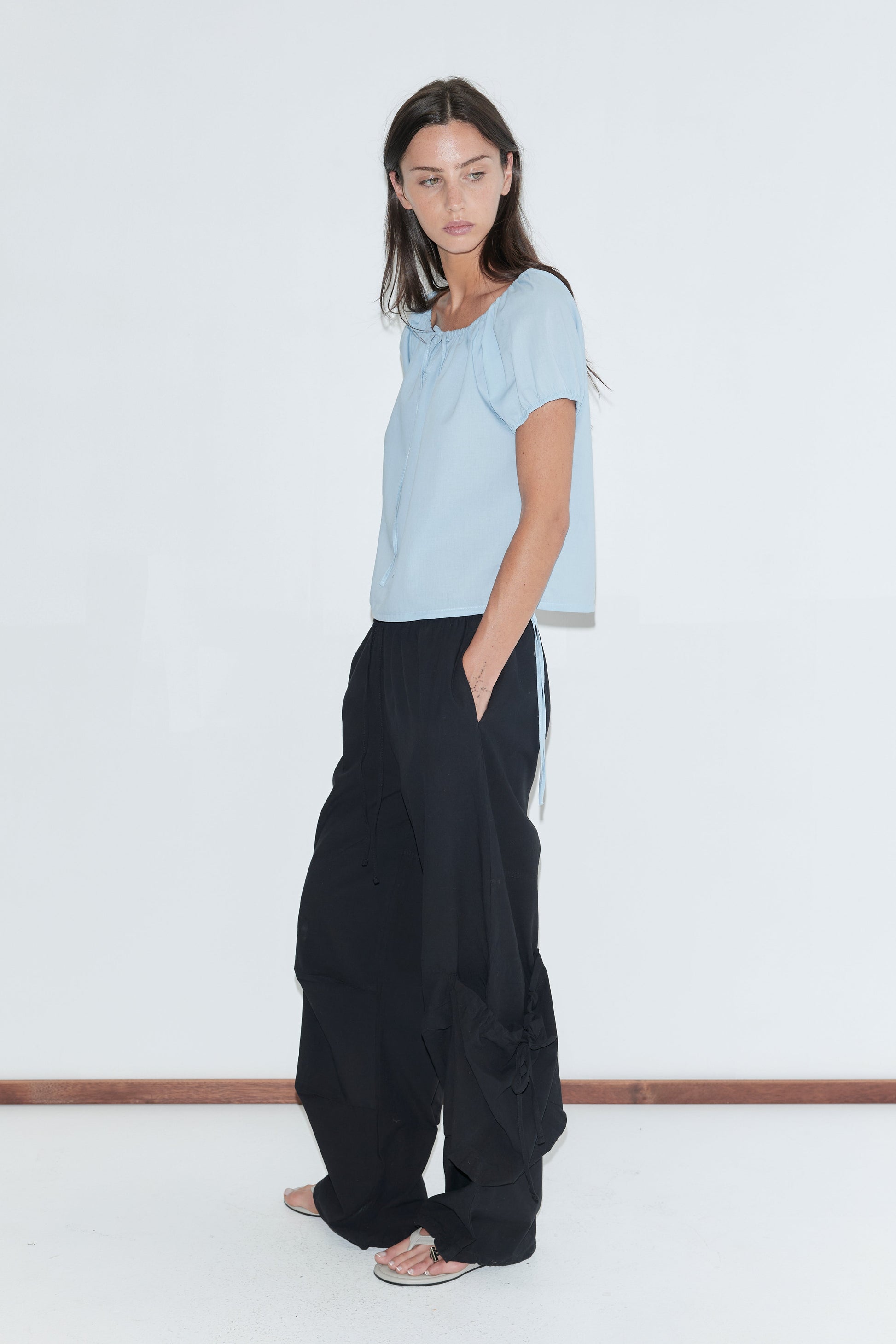 A woman with long dark hair stands in profile against a plain white background wearing the Drawcord Pocket Pant - Black by Deiji Studios, black wide-leg trousers with drawcord-gathered lower legs, with a pale blue top and grey flip-flops.