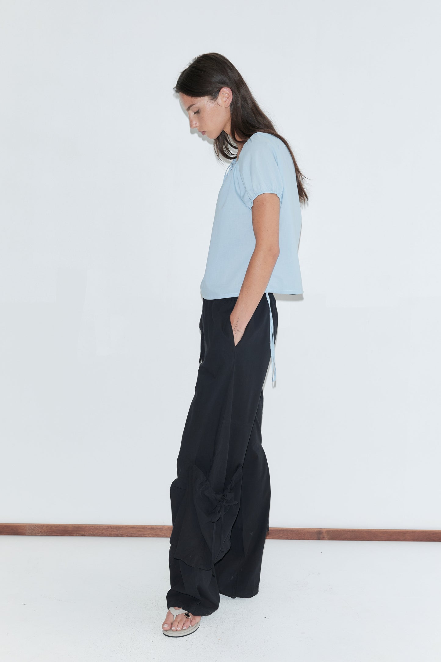 A woman with long dark hair stands in profile against a plain white background wearing the Drawcord Pocket Pant - Black by Deiji Studios, black wide-leg trousers, with a pale blue top and flip-flops.
