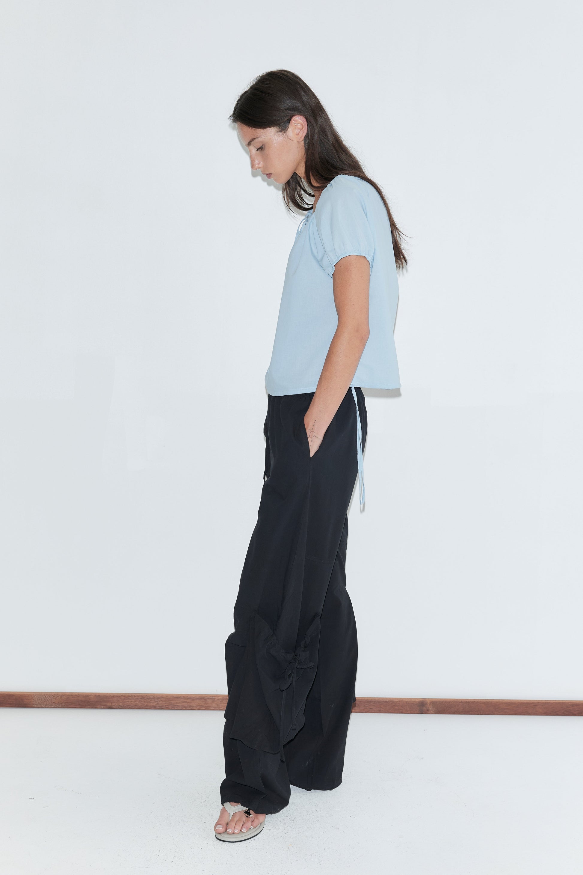 A woman with long dark hair stands in profile against a plain white background wearing the Drawcord Pocket Pant - Black by Deiji Studios, black wide-leg trousers, with a pale blue top and flip-flops.