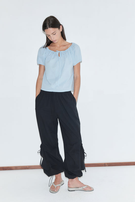A woman with long dark hair stands against a plain white background looking down, wearing the Drawcord Pocket Pant - Black by Deiji Studios, black wide-leg trousers with drawcord-gathered lower legs, with a pale blue tie top and flip-flops.