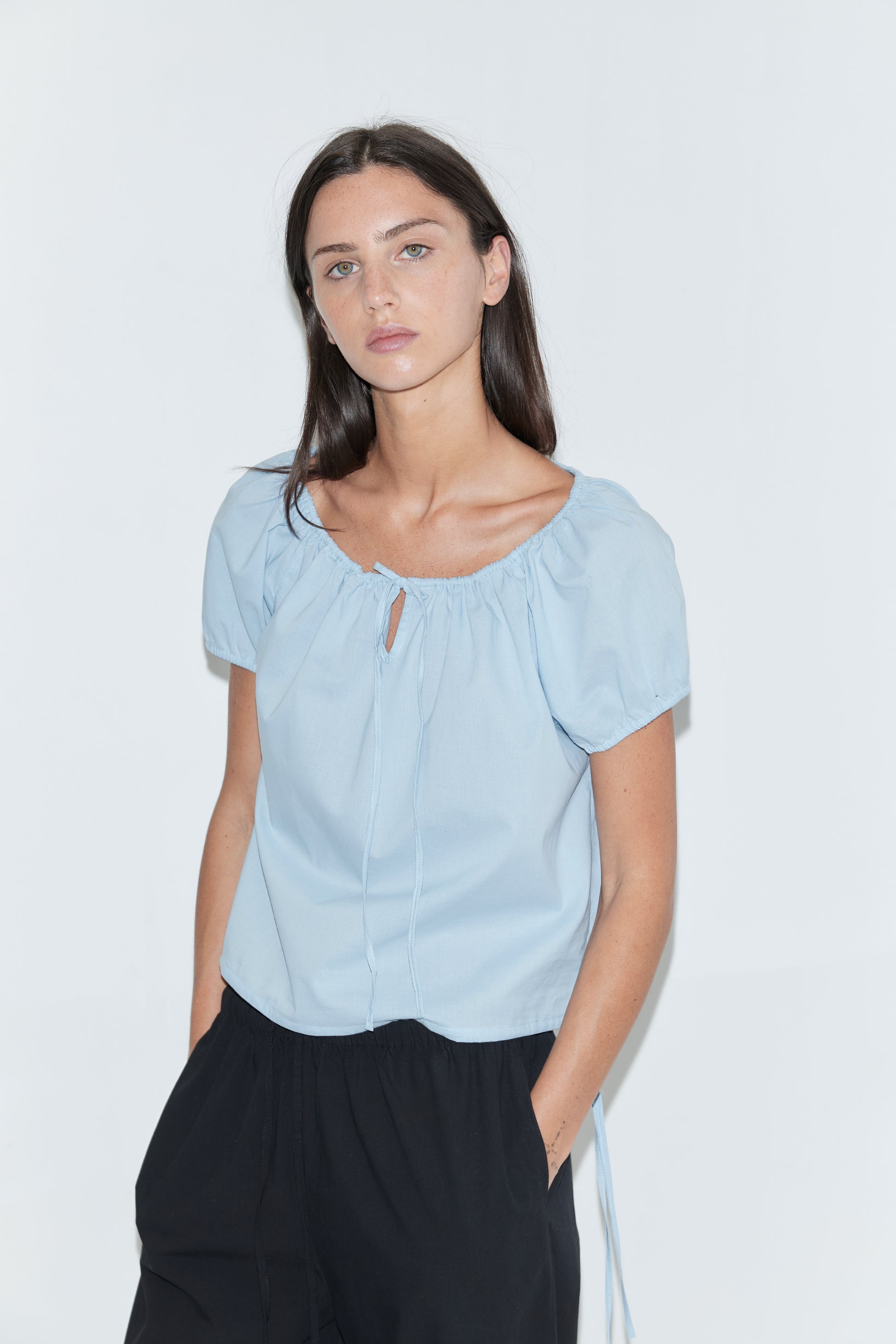 A woman with long dark hair stands against a plain white background wearing the Tie Top - Blue by Deiji Studios, a pale blue top with a gathered neckline, keyhole drawstring tie and puff sleeves, with black trousers.