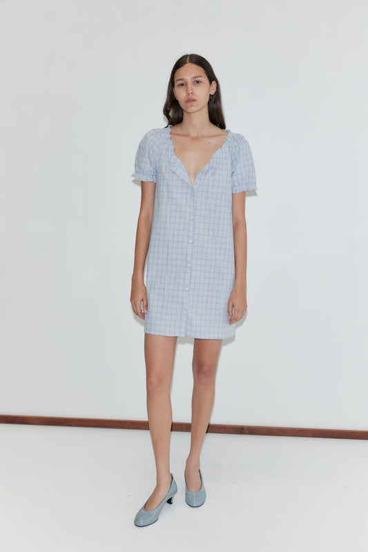 A woman with long dark hair wears the Buttoned Mini Dress - Powder Check and light blue heels, standing against a plain white background.