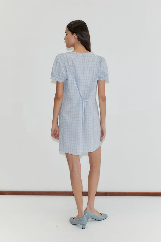 A woman with long brown hair stands facing away, wearing the Buttoned Mini Dress - Powder Check with a waist tie and light blue shoes, set against a plain white background.