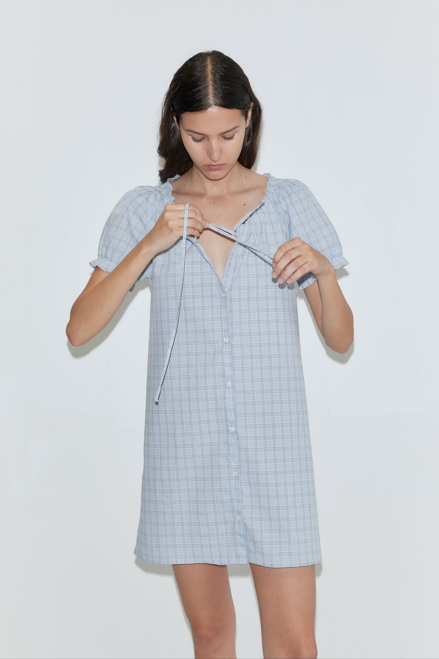 A woman with straight brown hair wears the Buttoned Mini Dress - Powder Check from Deiji Studios, tying the front neckline as she stands against a plain white background.